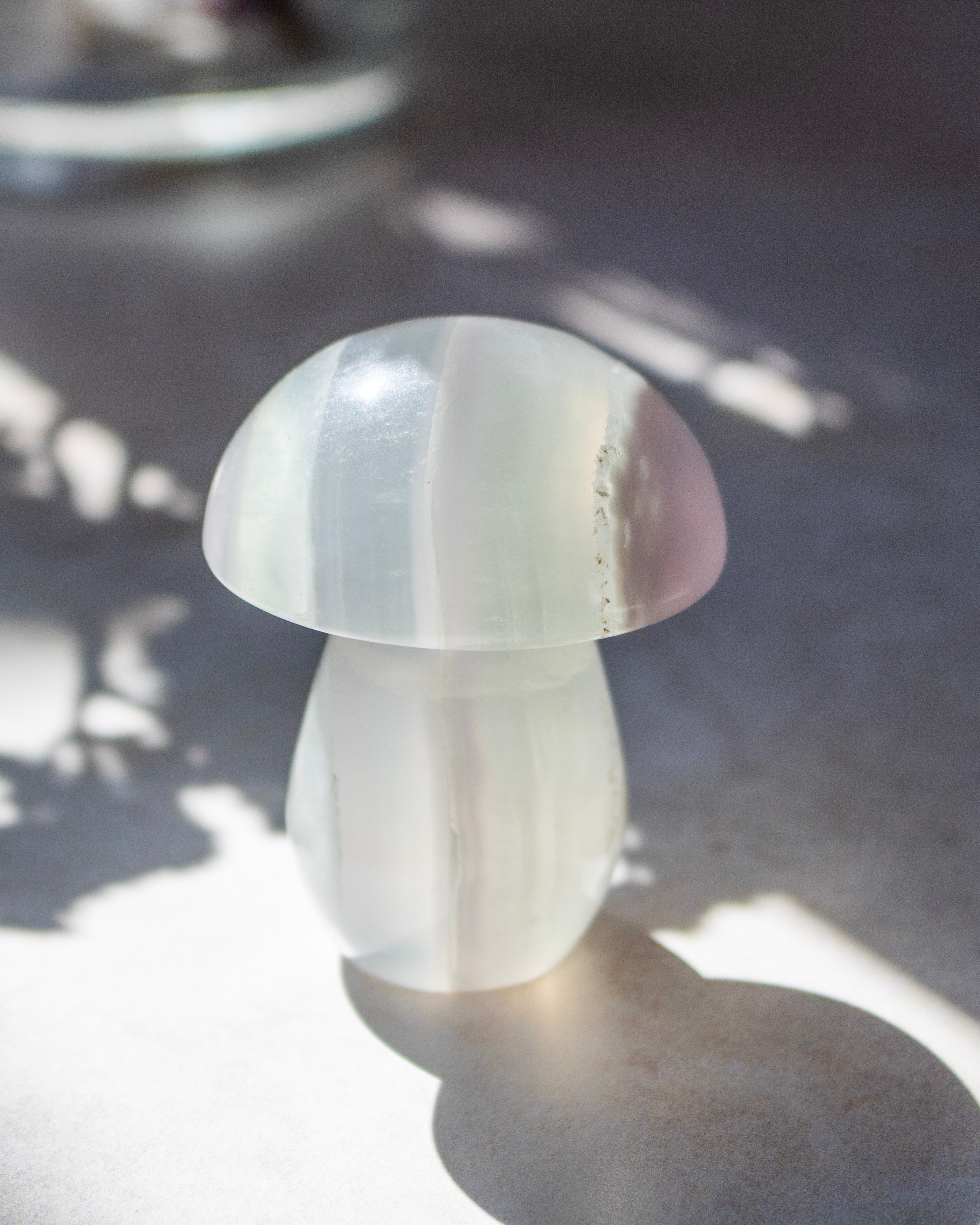 A small mushroom-shaped crystal sculpture made of milky translucent stone, photographed on a gray surface with dappled sunlight and shadows.