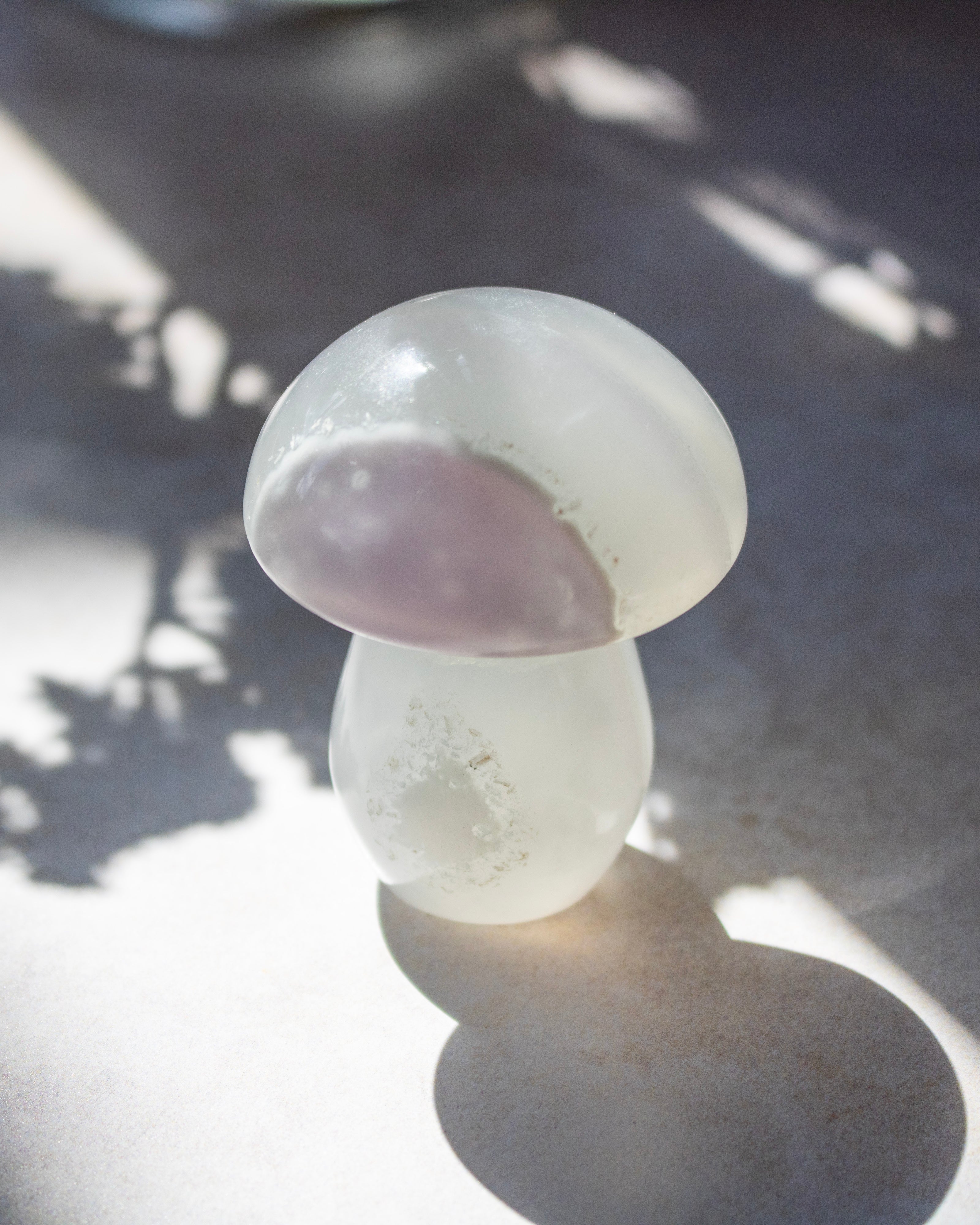 A mushroom-shaped crystal healing stone with a translucent white body and a pale lavender-purple cap, resting on a gray surface with dappled sunlight and leaf shadows.