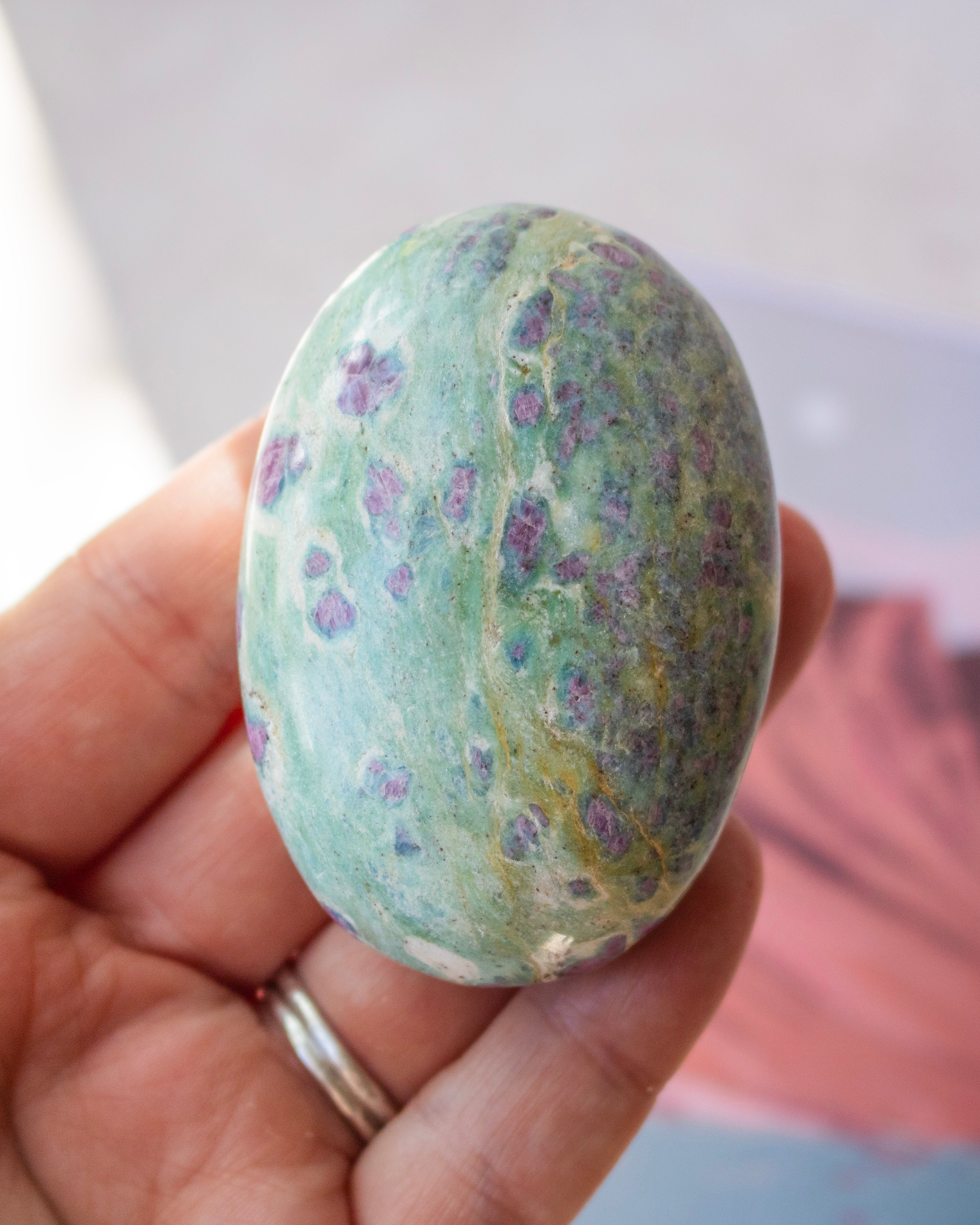A hand holding a polished oval stone with a mottled green and blue surface and scattered purple crystal formations, wearing a silver ring.