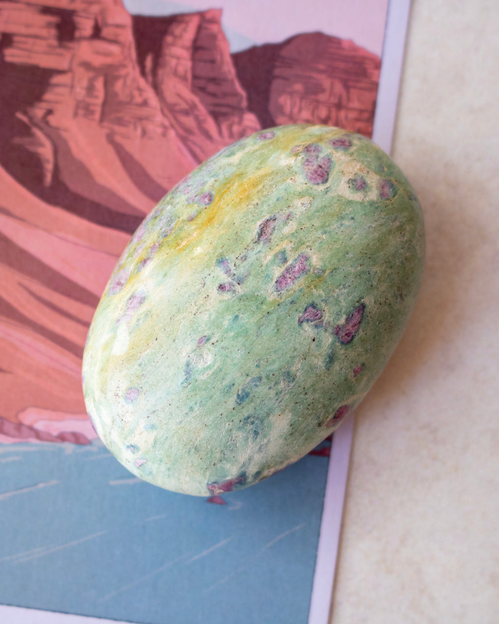 A green stone or polished rock with purple and cream marbling, resting on an illustrated card depicting red rock canyons.