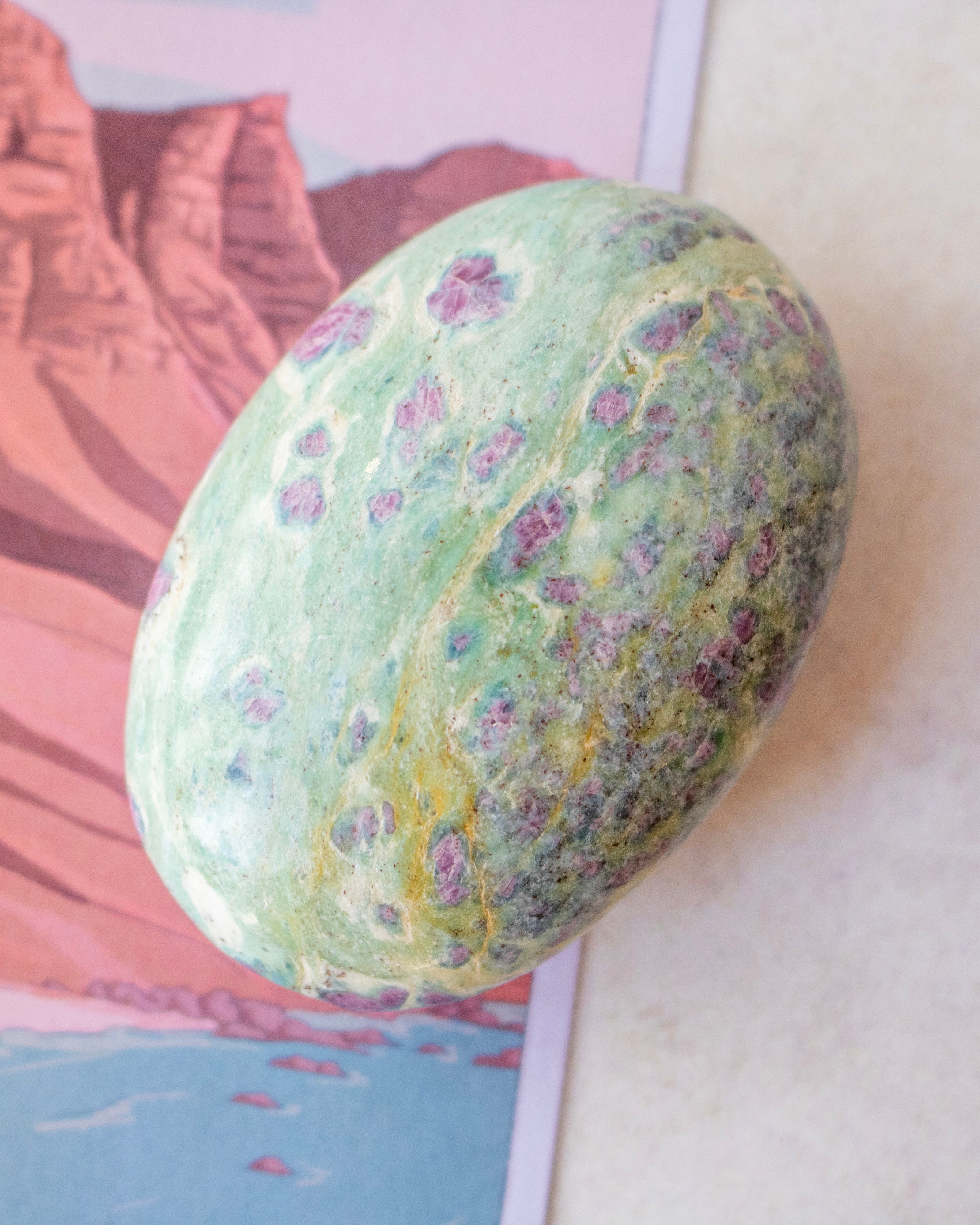 A polished green stone with purple and yellow mineral inclusions, resting on a printed card with a desert landscape illustration.