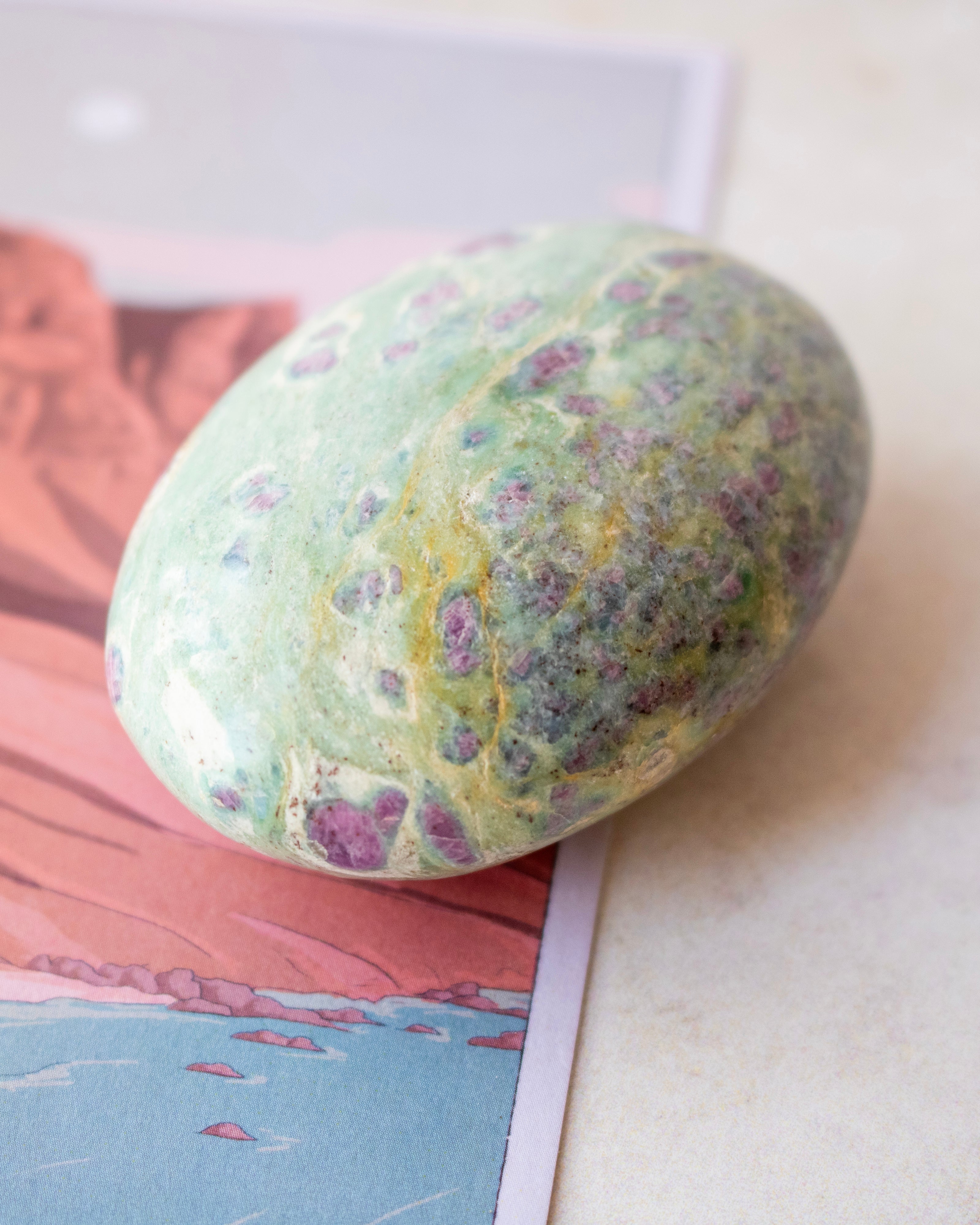 A polished green stone with purple and yellow marbling, resting on a printed card against a light neutral background.