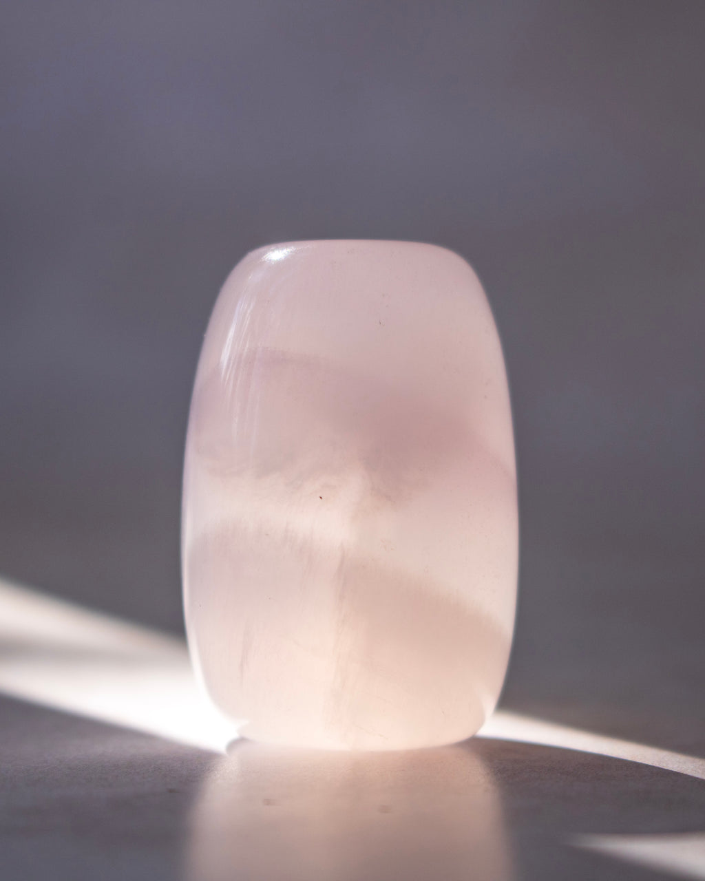 A polished pale pink stone or crystal holder with rounded edges, standing upright on a reflective surface against a soft gray background with sunlight casting shadows.