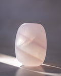 A polished pale pink stone or crystal with rounded edges, standing upright on a dark surface against a soft gray background with sunlight casting a shadow.
