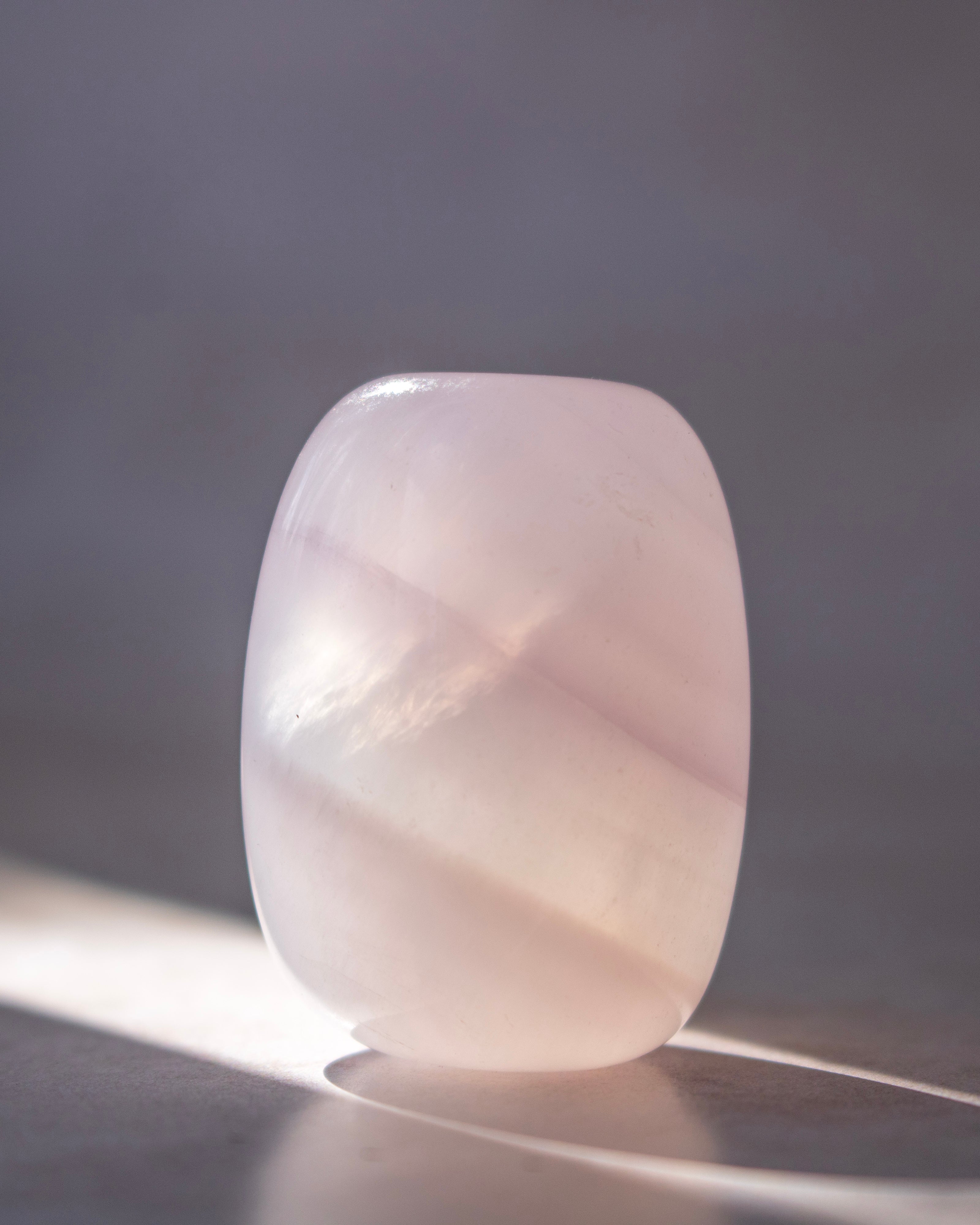 A polished pale pink stone or crystal with rounded edges, standing upright on a dark surface against a soft gray background with sunlight casting a shadow.