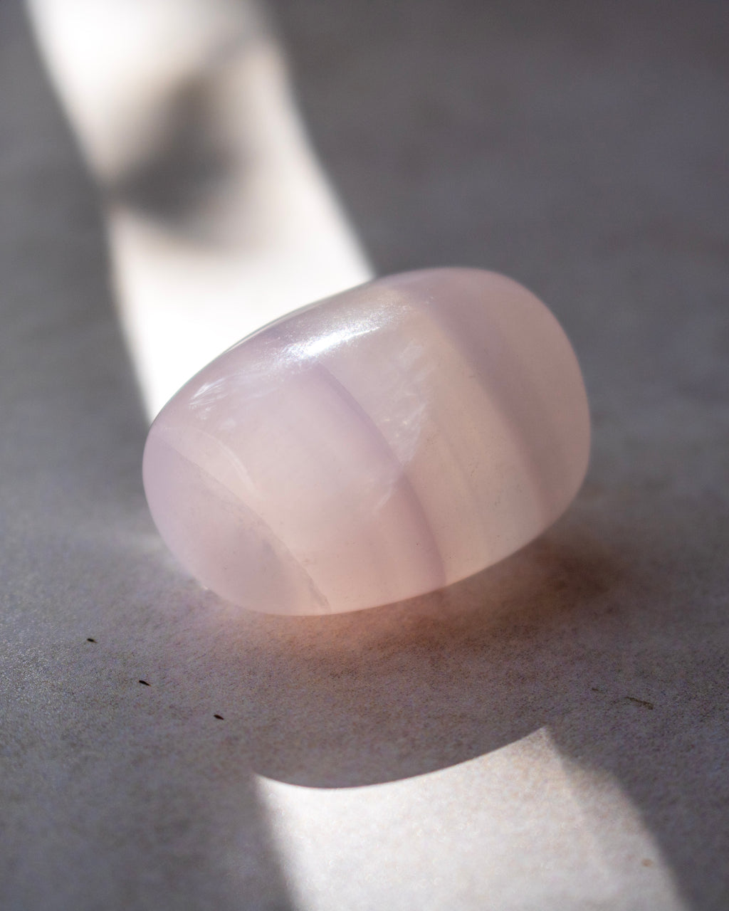 A smooth, polished pale pink stone or gemstone with a rounded teardrop shape, resting on a textured gray surface under dramatic, directional light that casts a soft shadow.