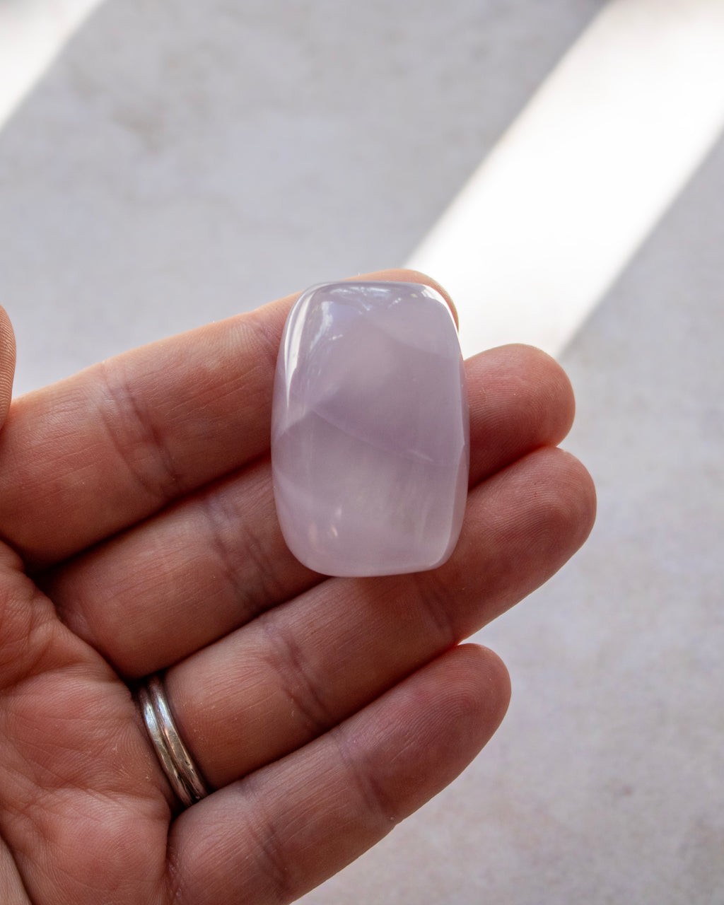 A hand holding a smooth, polished pale pink stone cabochon against a light gray concrete background.