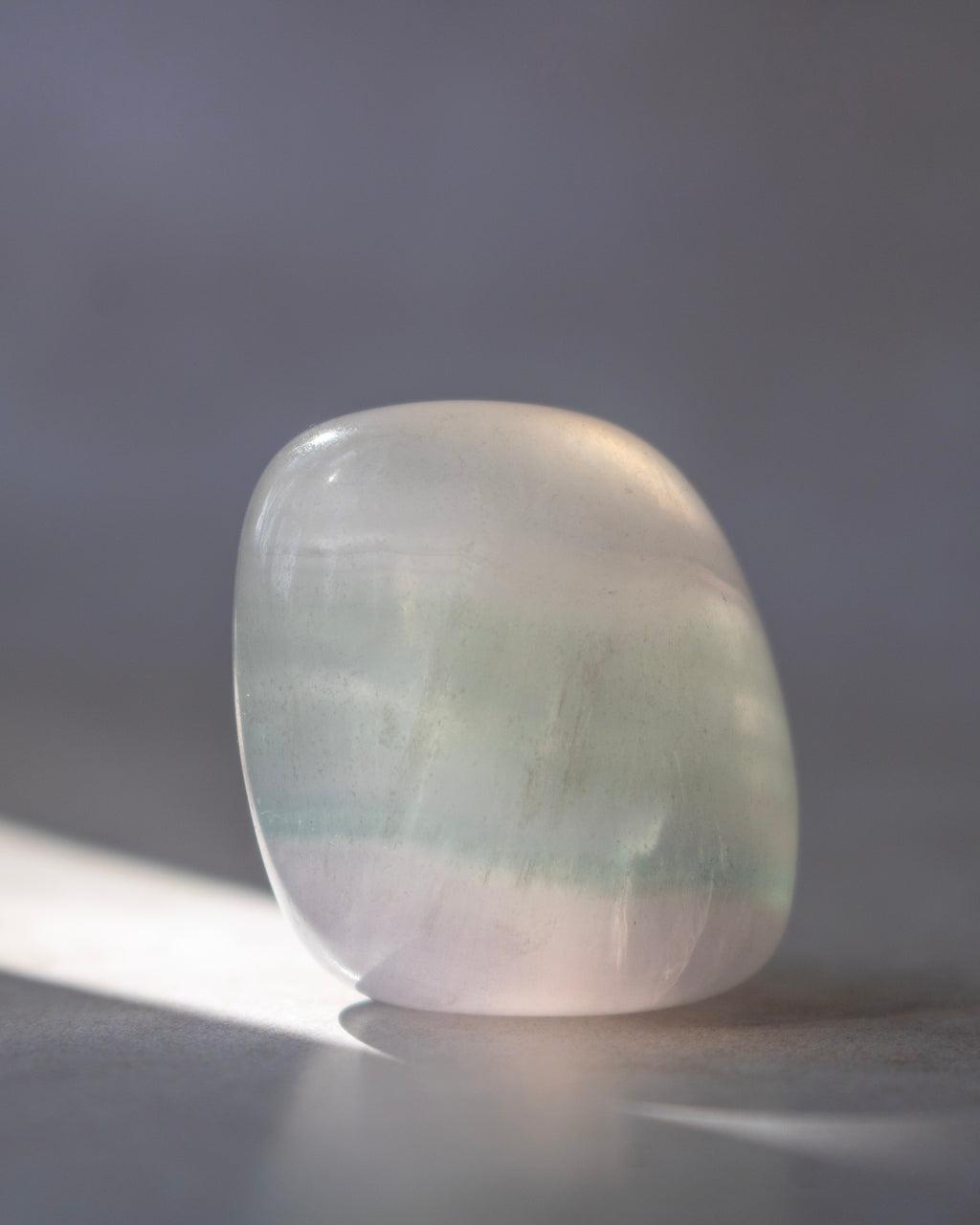 A polished tumbled stone with soft pale green and lavender-pink tones, photographed on a neutral gray surface with a soft shadow.