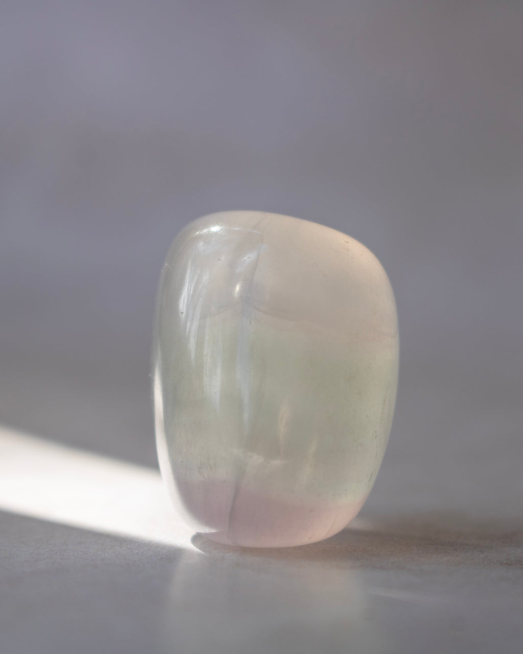 A polished, rounded tumbled stone with a soft pearlescent finish, showing subtle pastel gradients of pale green, pink, and cream under warm directional light.