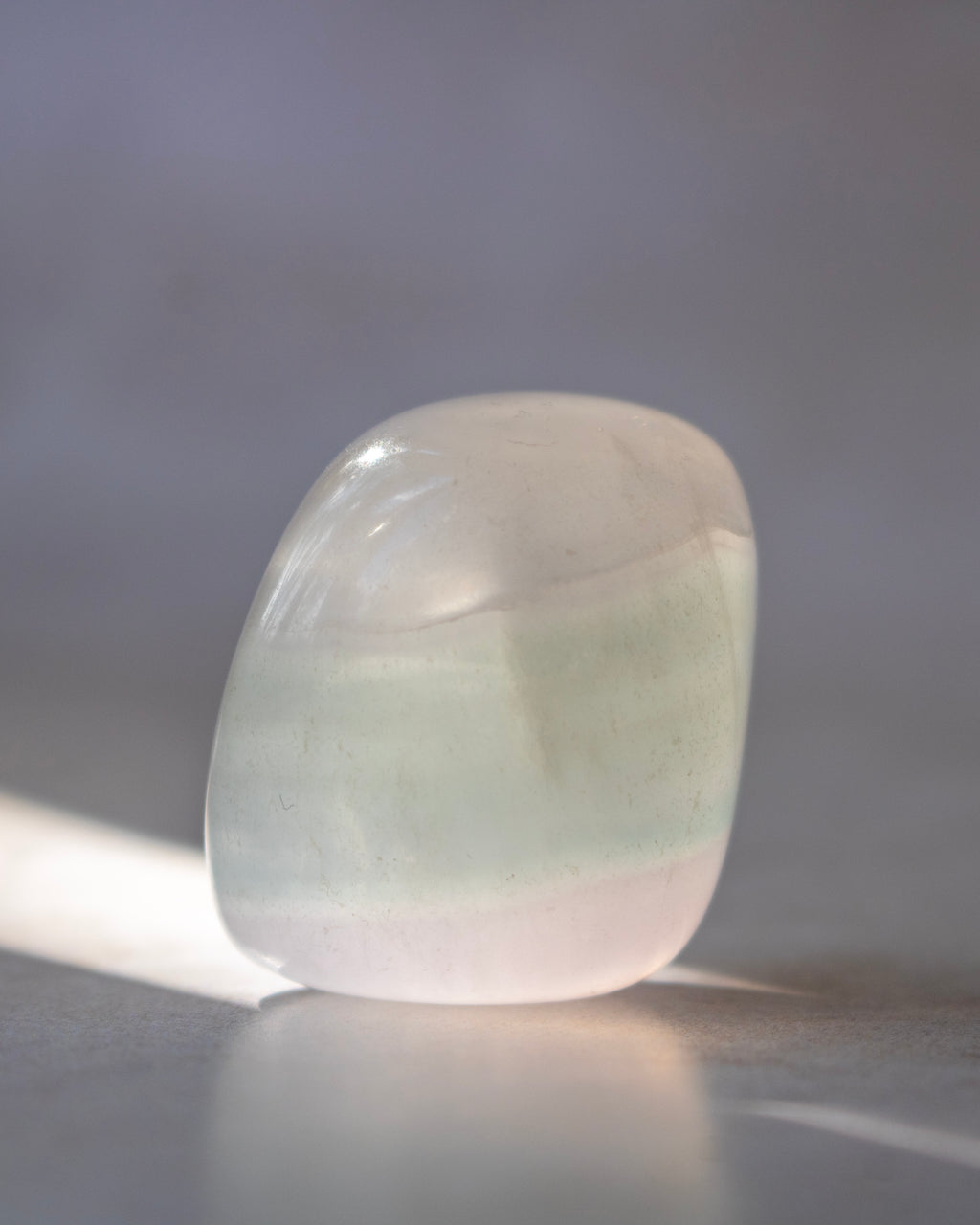 A polished, rounded tumbled stone with a soft pale green and white coloration, resting on a reflective surface against a soft gray background.