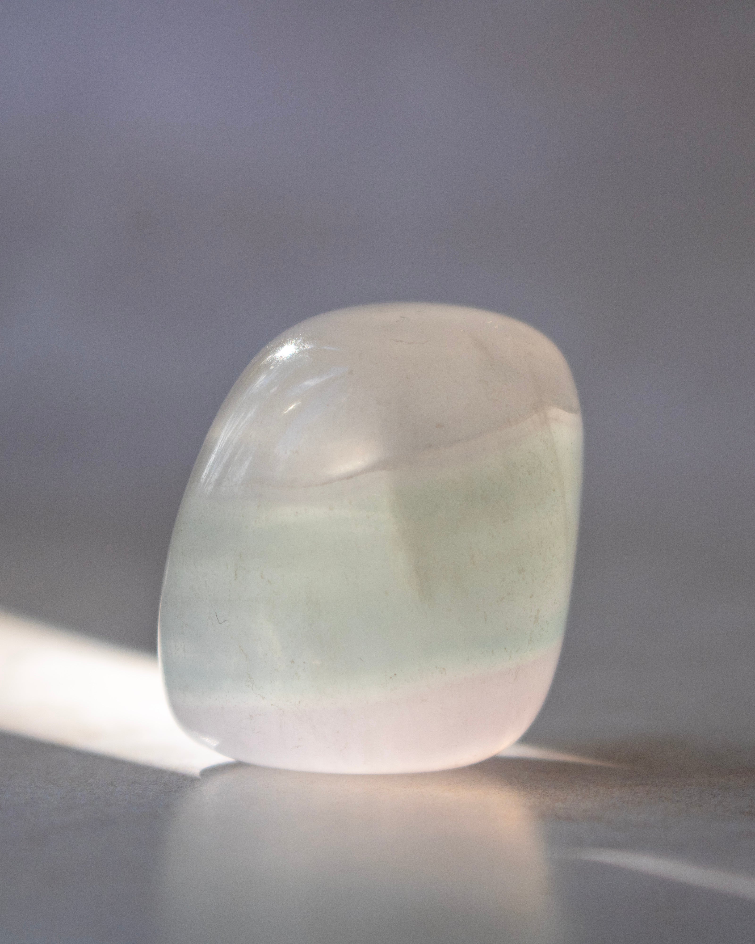 A polished, rounded tumbled stone with a soft pale green and white coloration, resting on a reflective surface against a soft gray background.