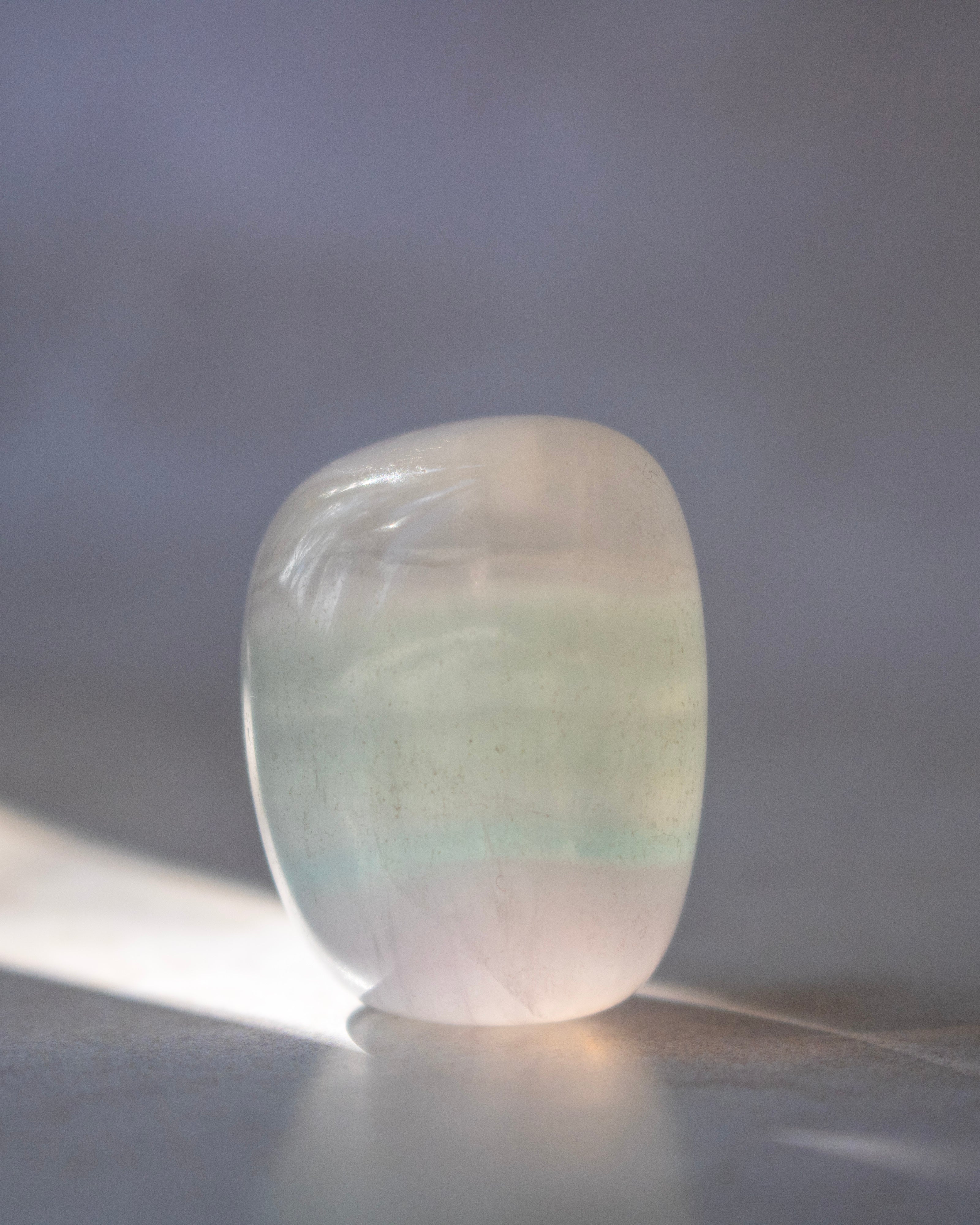 A polished tumbled stone crystal with a rounded rectangular shape, featuring a soft iridescent color shift from pale green to light pink, resting on a reflective surface with a soft shadow.