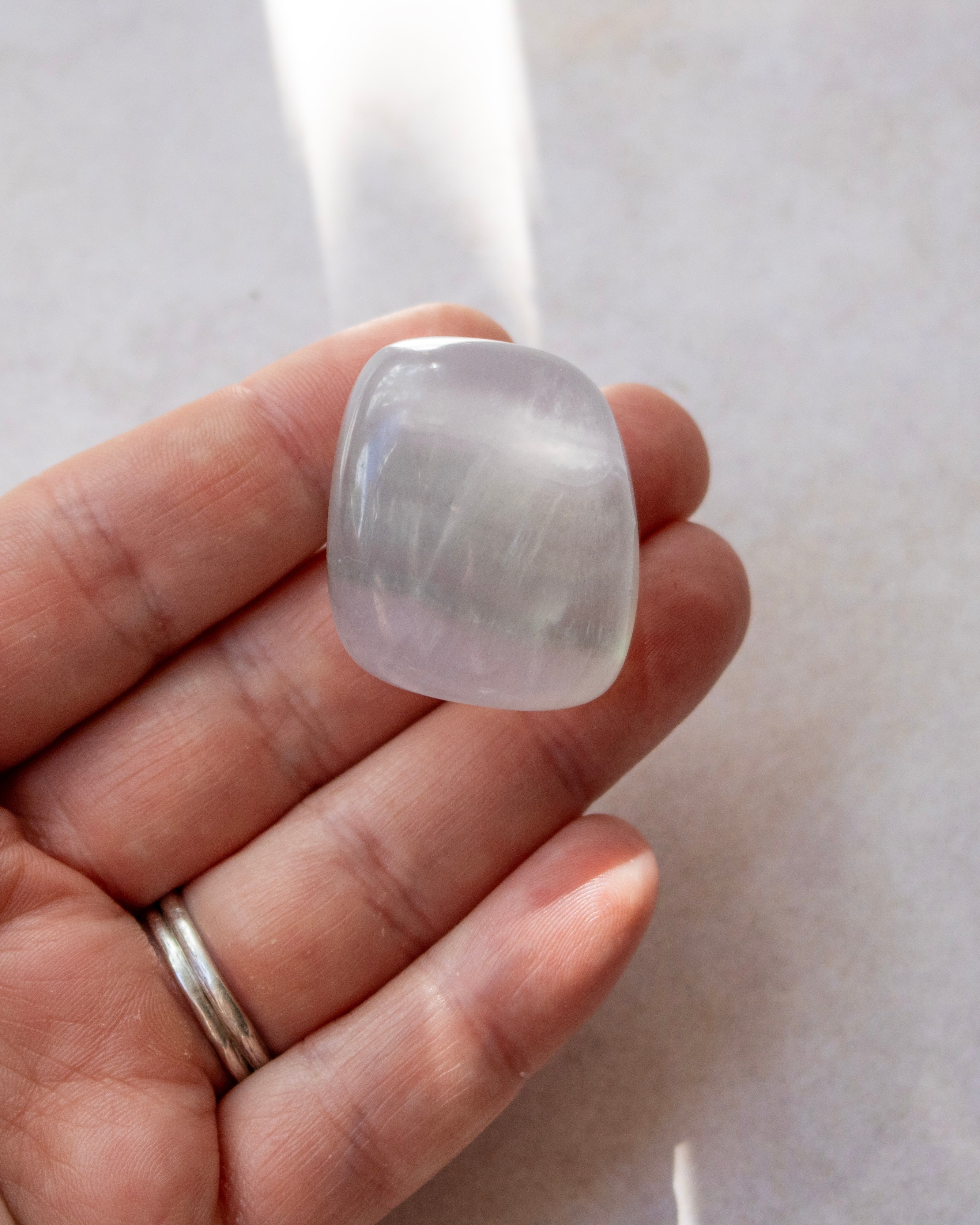 A hand holding a smooth, rounded translucent stone with a pale lavender-gray hue and fine crystalline texture, set against a light neutral background.