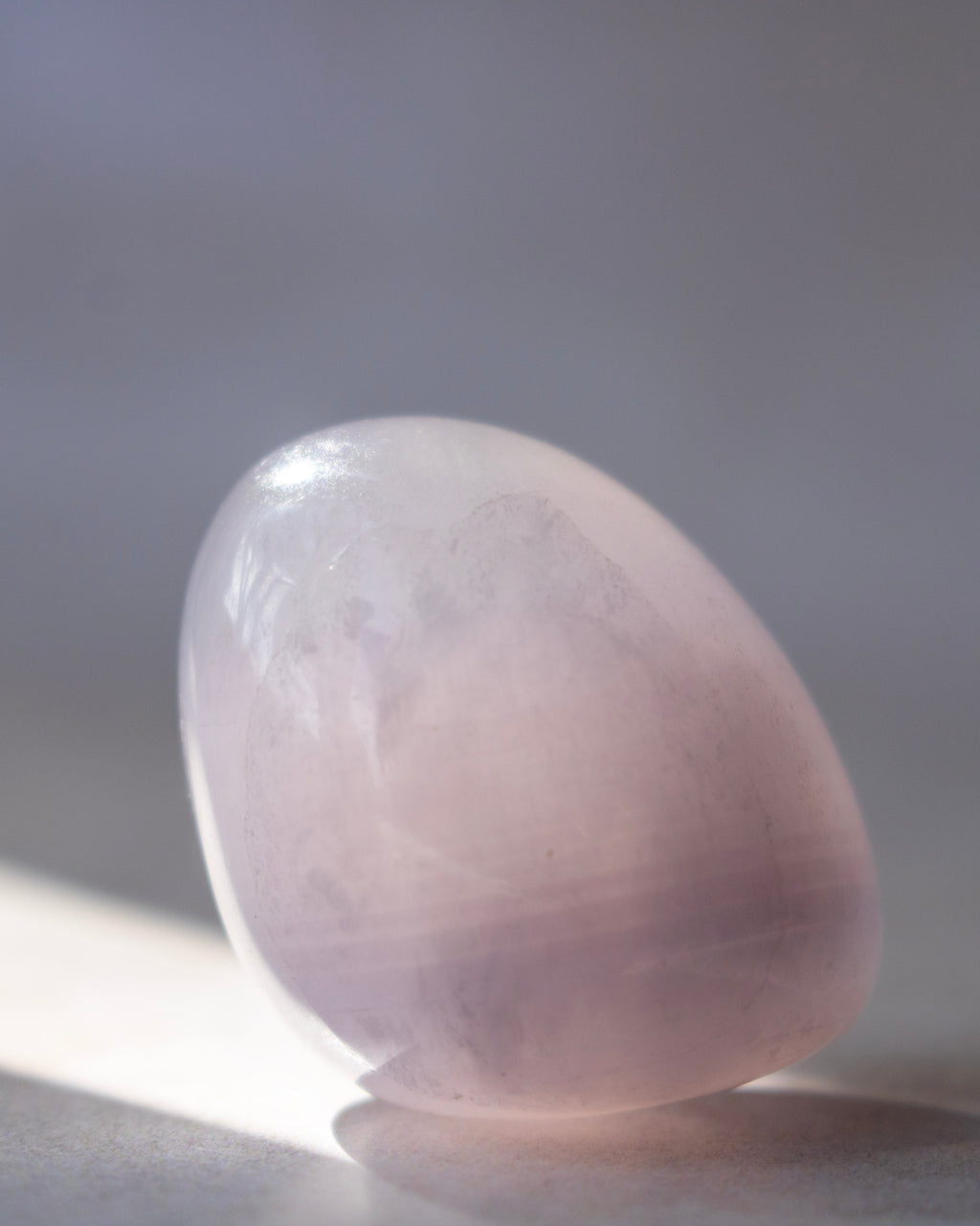 A polished crystal tumbled into an oval shape, resting on a neutral gray surface with soft sunlight casting a shadow.