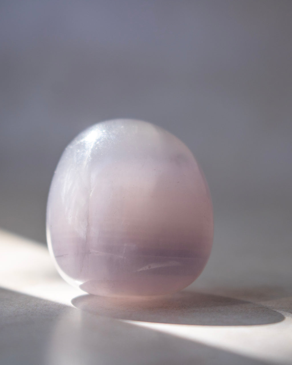A smooth, polished oval stone with a soft lavender-pink hue, resting on a light surface with a soft shadow under direct sunlight.
