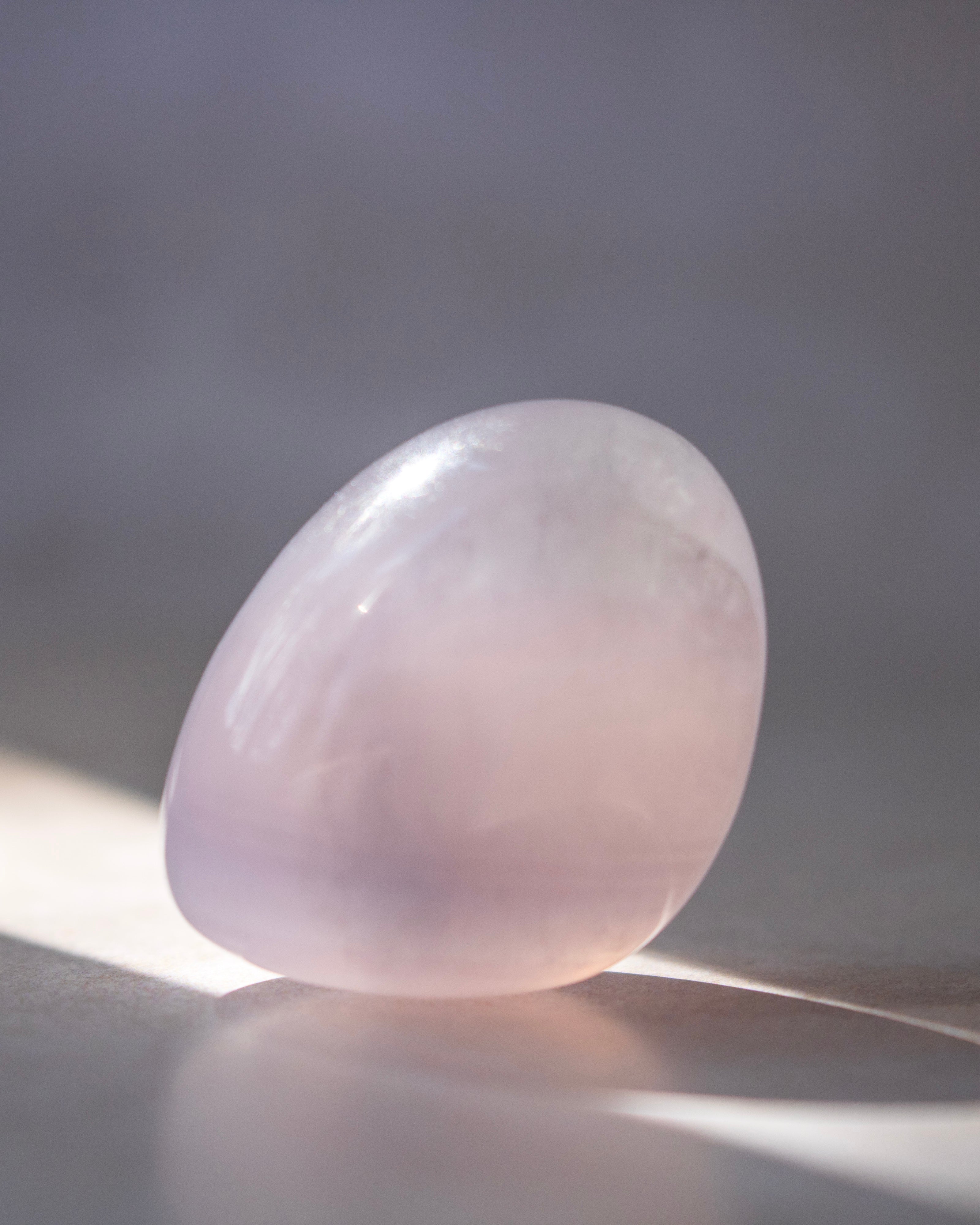 A smooth, polished rose quartz crystal sphere resting on a reflective surface with soft sunlight and shadows.