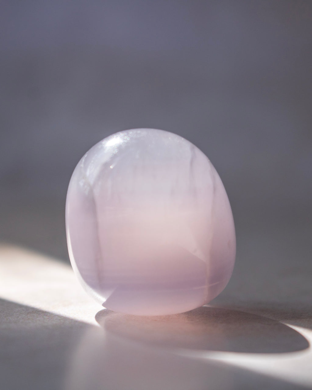A smooth, polished oval stone in a pale pinkish-lavender hue, standing on a surface with soft sunlight and shadow against a neutral gray background.