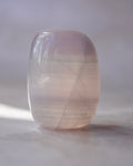 A polished crystal tumbler with smooth rounded edges, showing natural internal inclusions and a soft pink color, resting on a neutral surface.