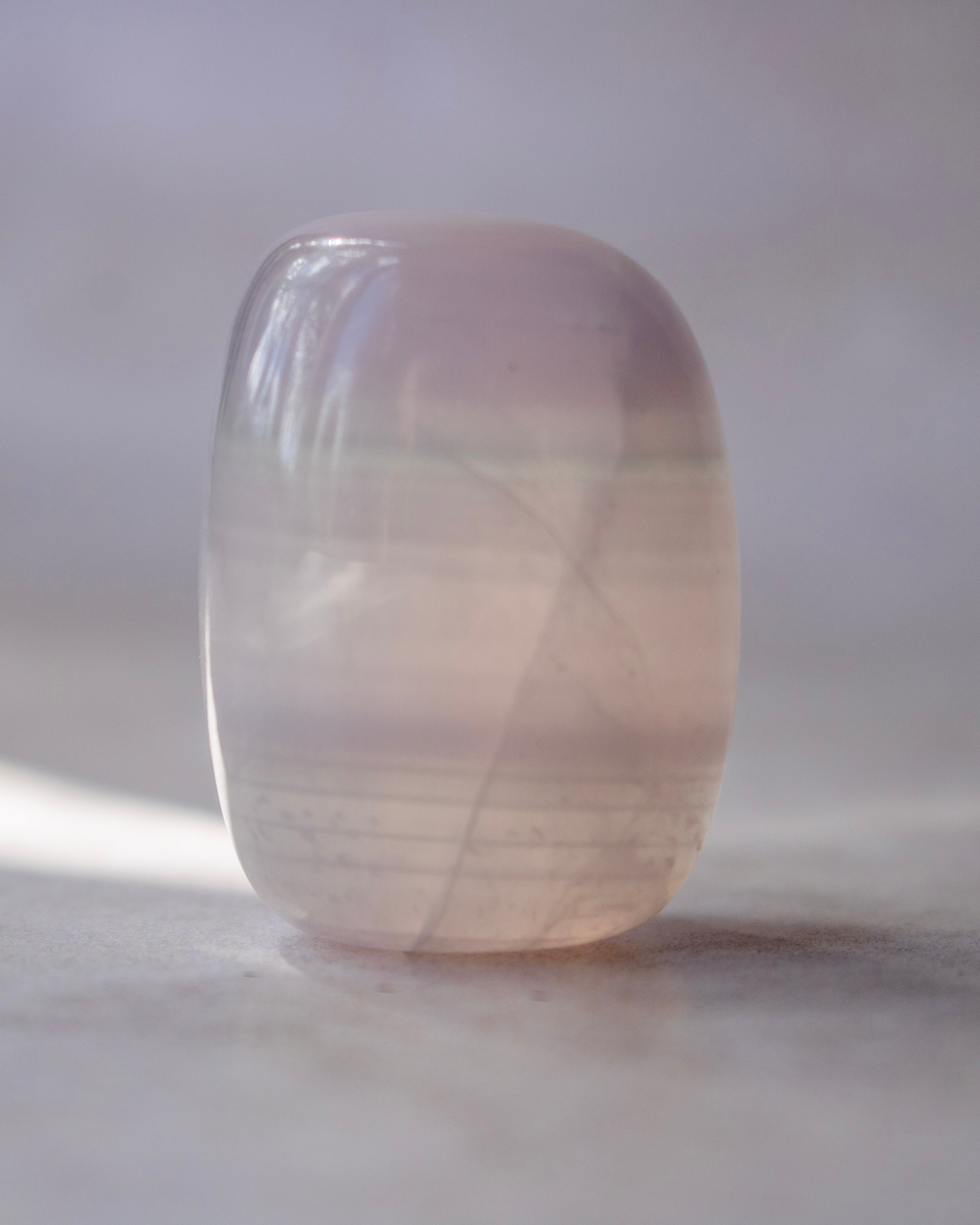 A polished crystal tumbler with smooth rounded edges, showing natural internal inclusions and a soft pink color, resting on a neutral surface.