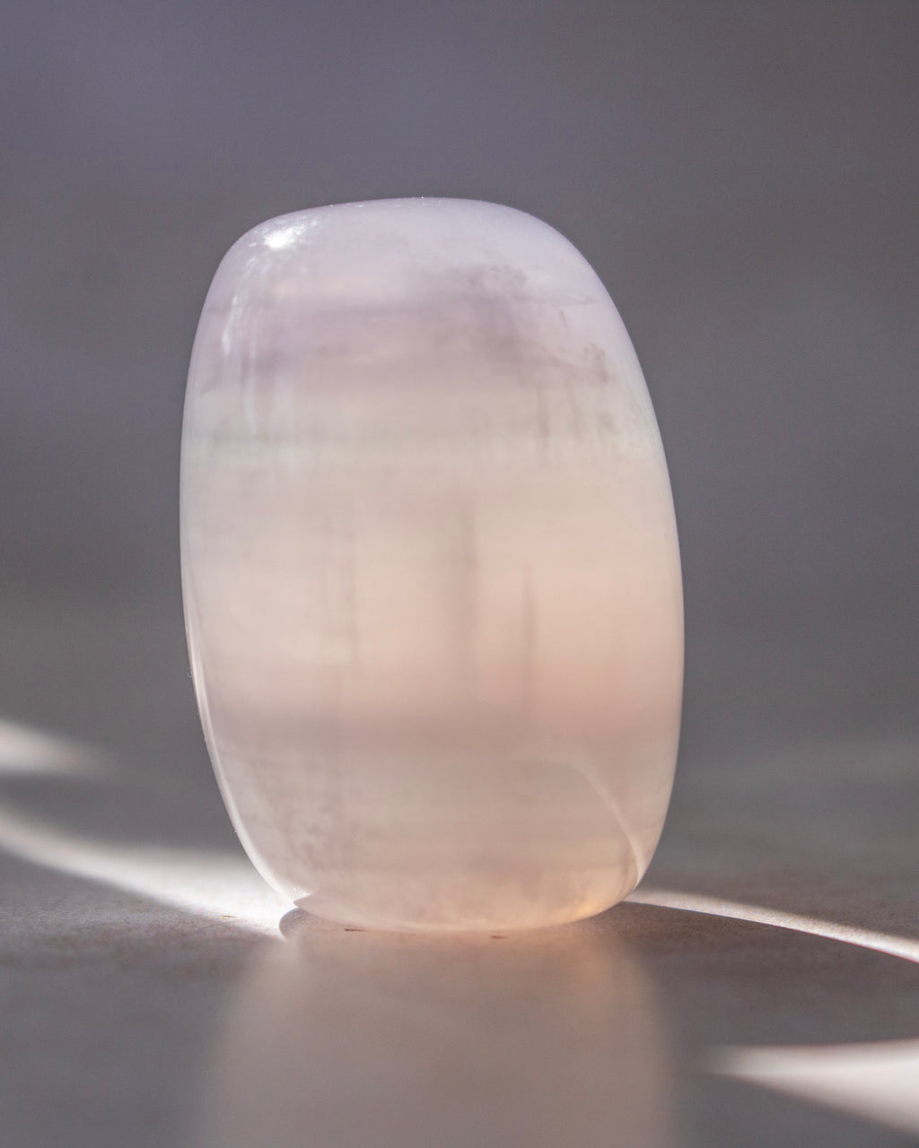 A smooth, rounded translucent stone or crystal sculpture in a pale blush pink color, standing on a reflective surface against a soft gray background.