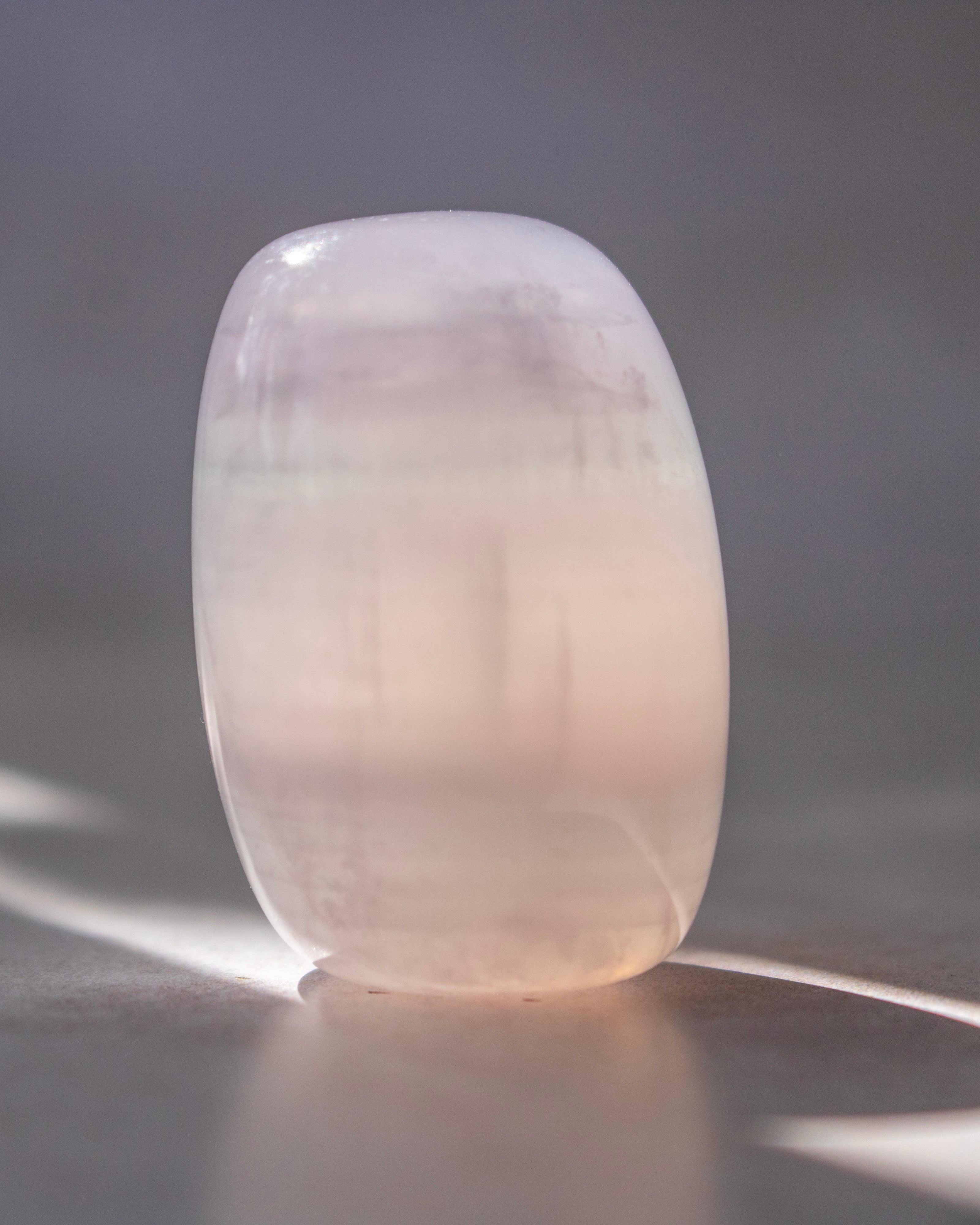 A smooth, rounded translucent stone or crystal sculpture in a pale blush pink color, standing on a reflective surface against a soft gray background.