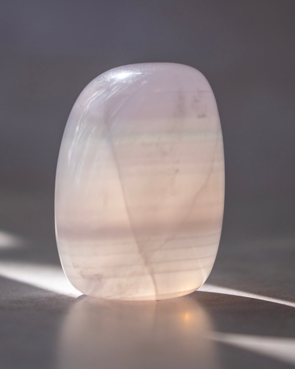 A polished, rounded translucent stone tumbler with soft pink and white tones, standing on a reflective surface with dappled sunlight casting shadows.