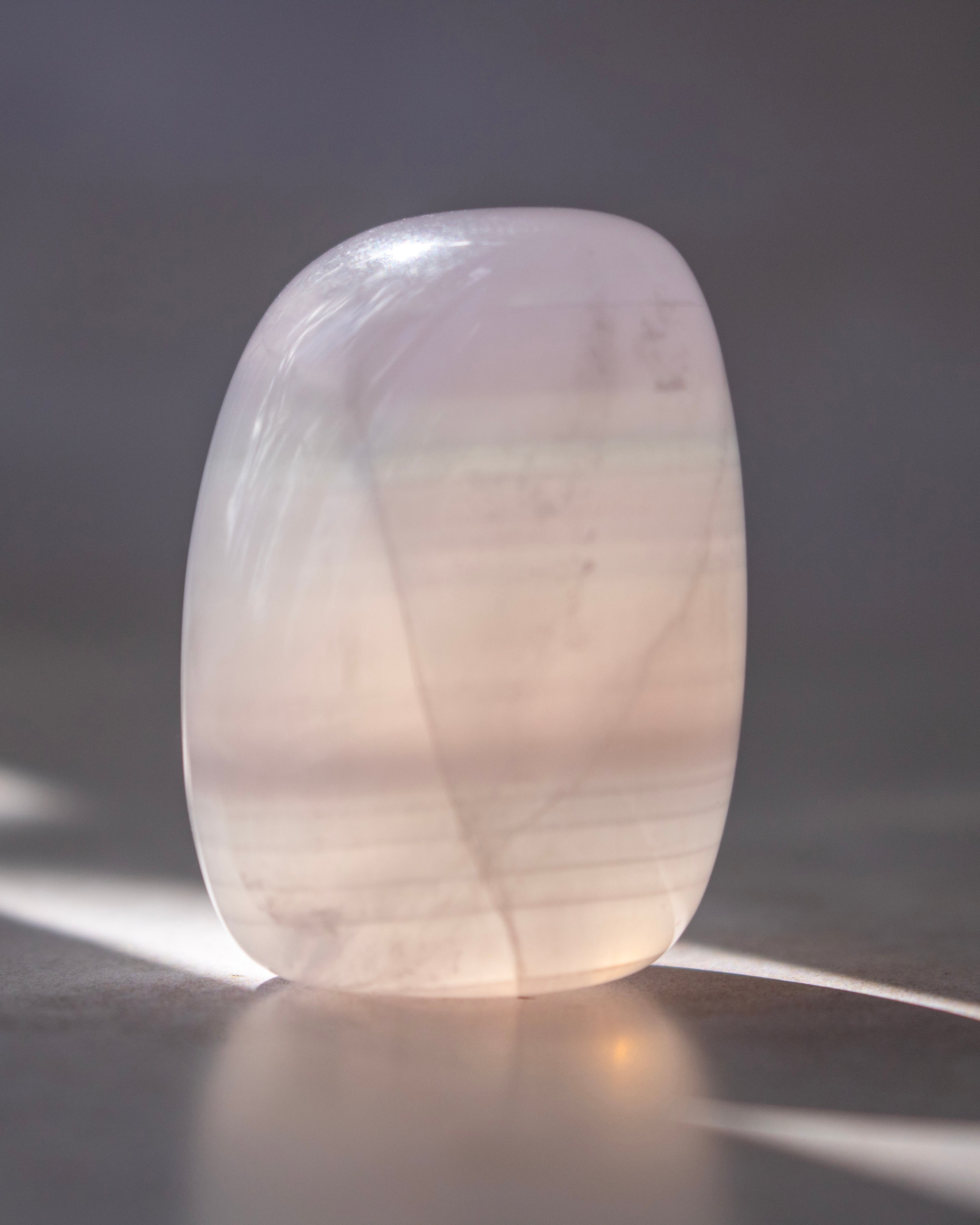 A polished, rounded translucent stone tumbler with soft pink and white tones, standing on a reflective surface with dappled sunlight casting shadows.