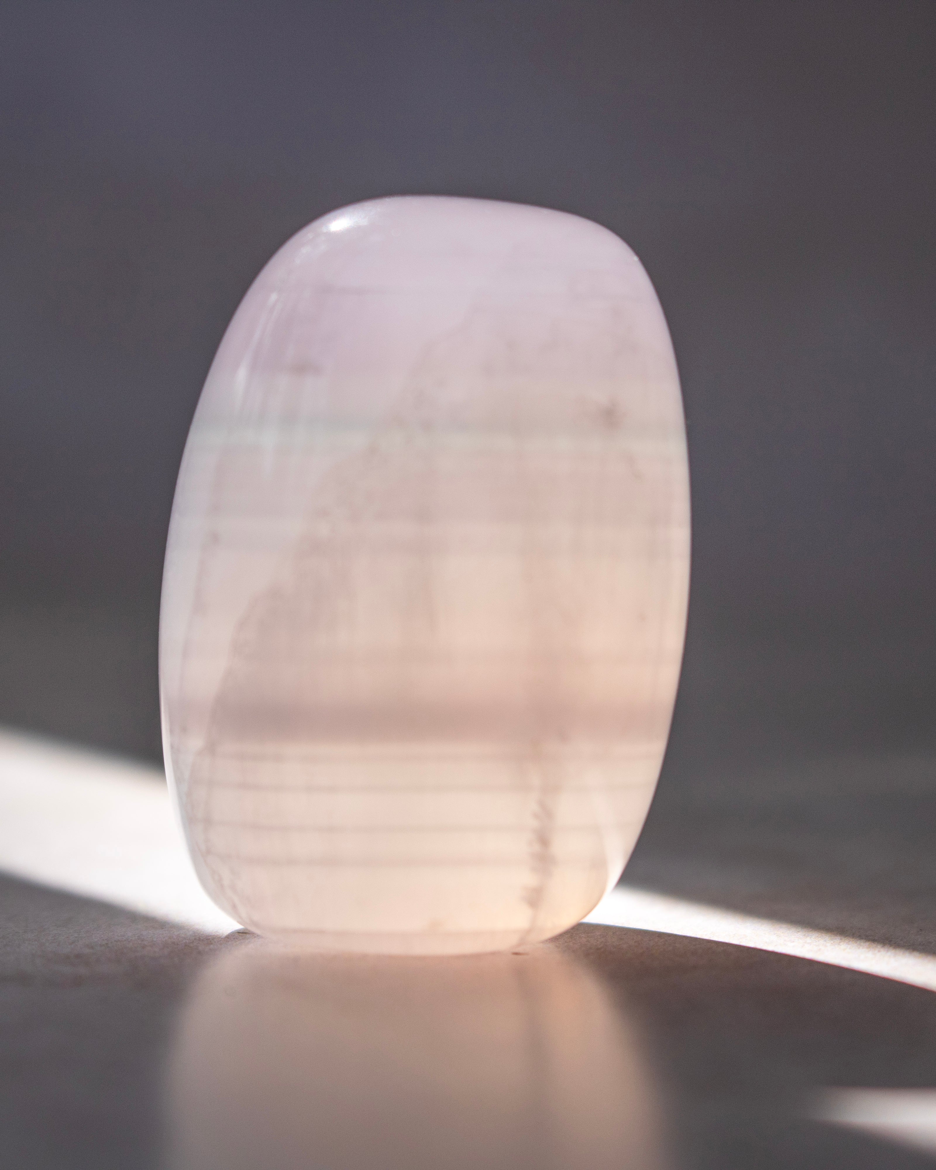 A polished pale pink stone tumbler with a smooth, rounded rectangular shape, standing on a dark reflective surface with soft light reflecting off it.