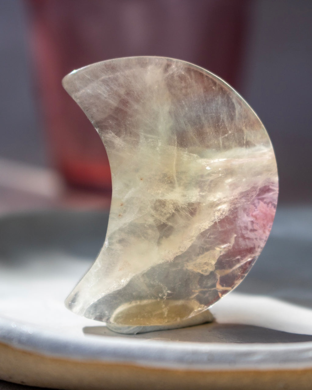 A crescent-shaped clear crystal stone with natural inclusions and a pinkish-purple section, displayed on a small stand on a ceramic surface.