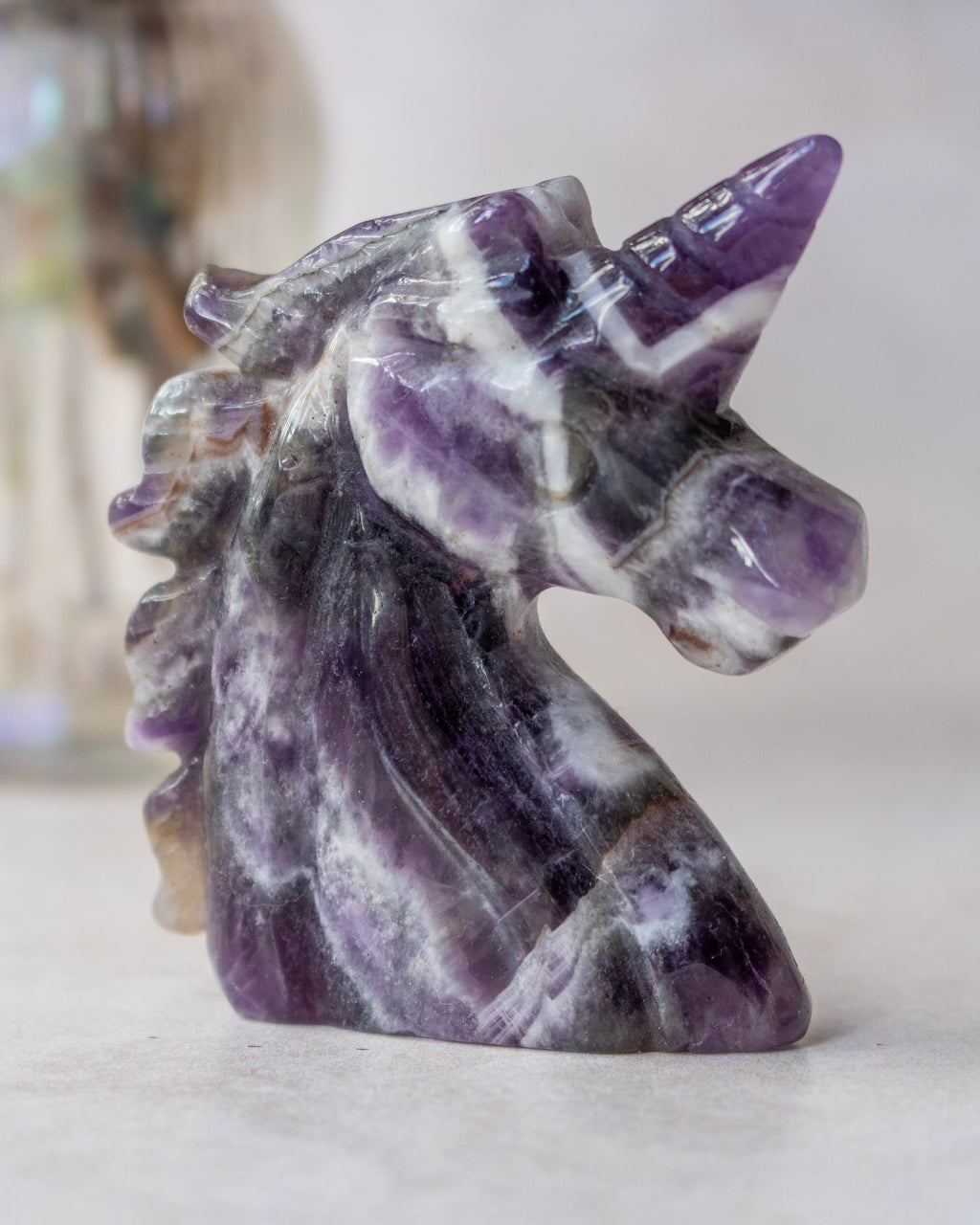 A polished carved unicorn head sculpture made from purple and white marbled stone, shown standing on a light surface against a soft neutral background.