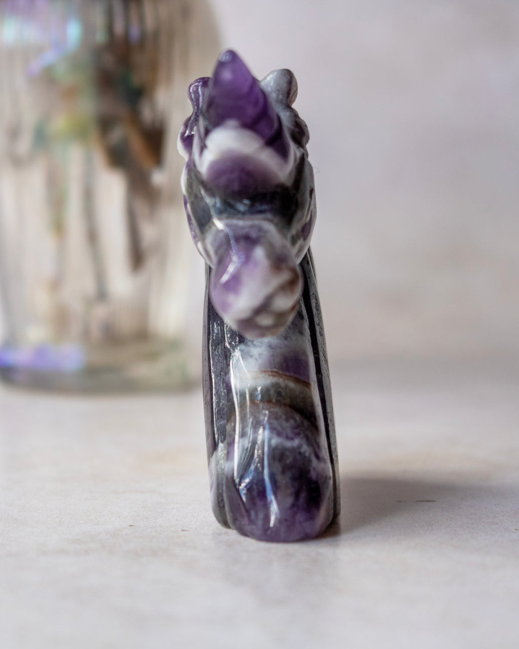 A small carved amethyst and white stone figurine, shaped like a stylized animal head or creature, standing upright on a light surface with a softly blurred glass container in the background.