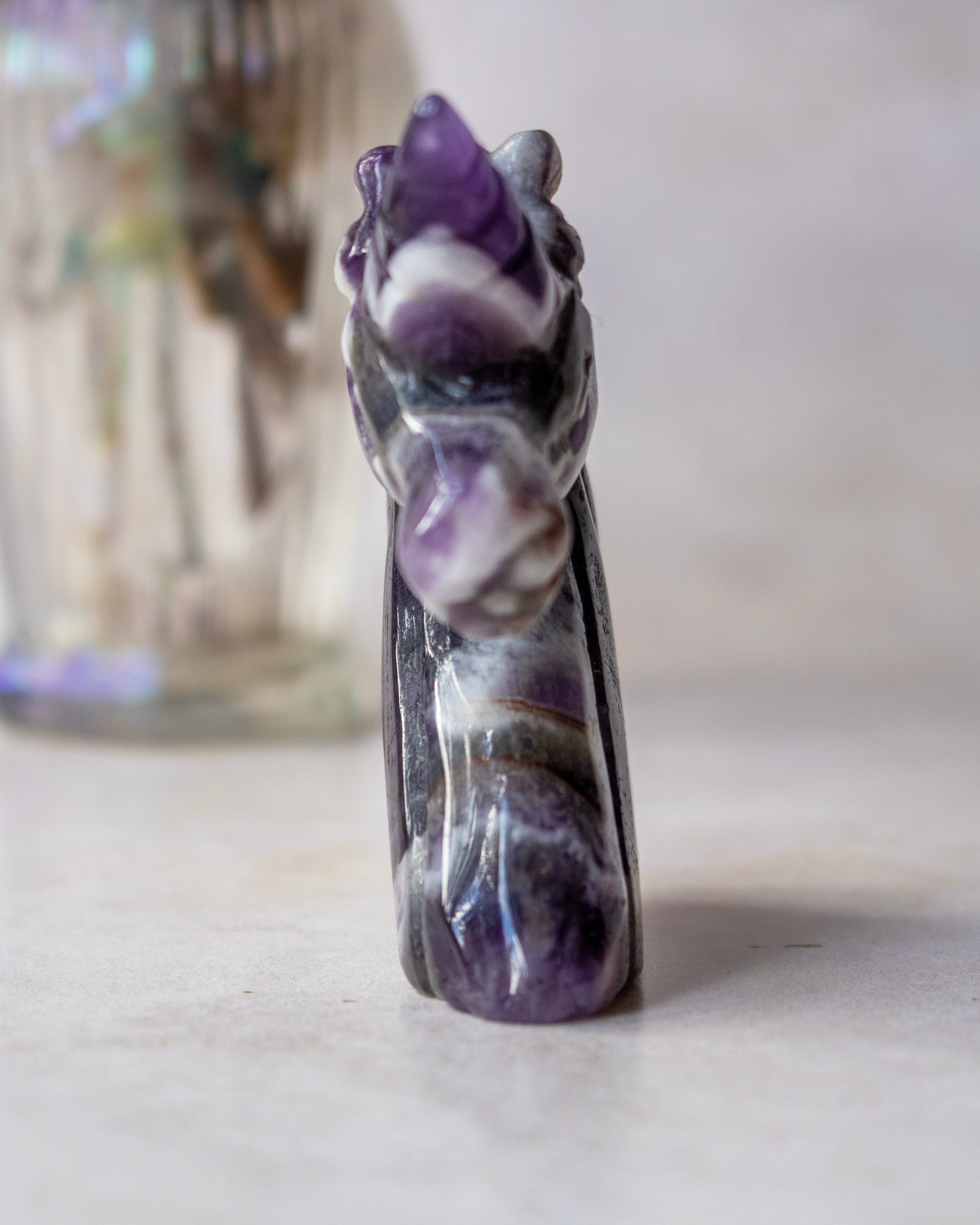 A small carved amethyst and white stone figurine, shaped like a stylized animal head or creature, standing upright on a light surface with a softly blurred glass container in the background.