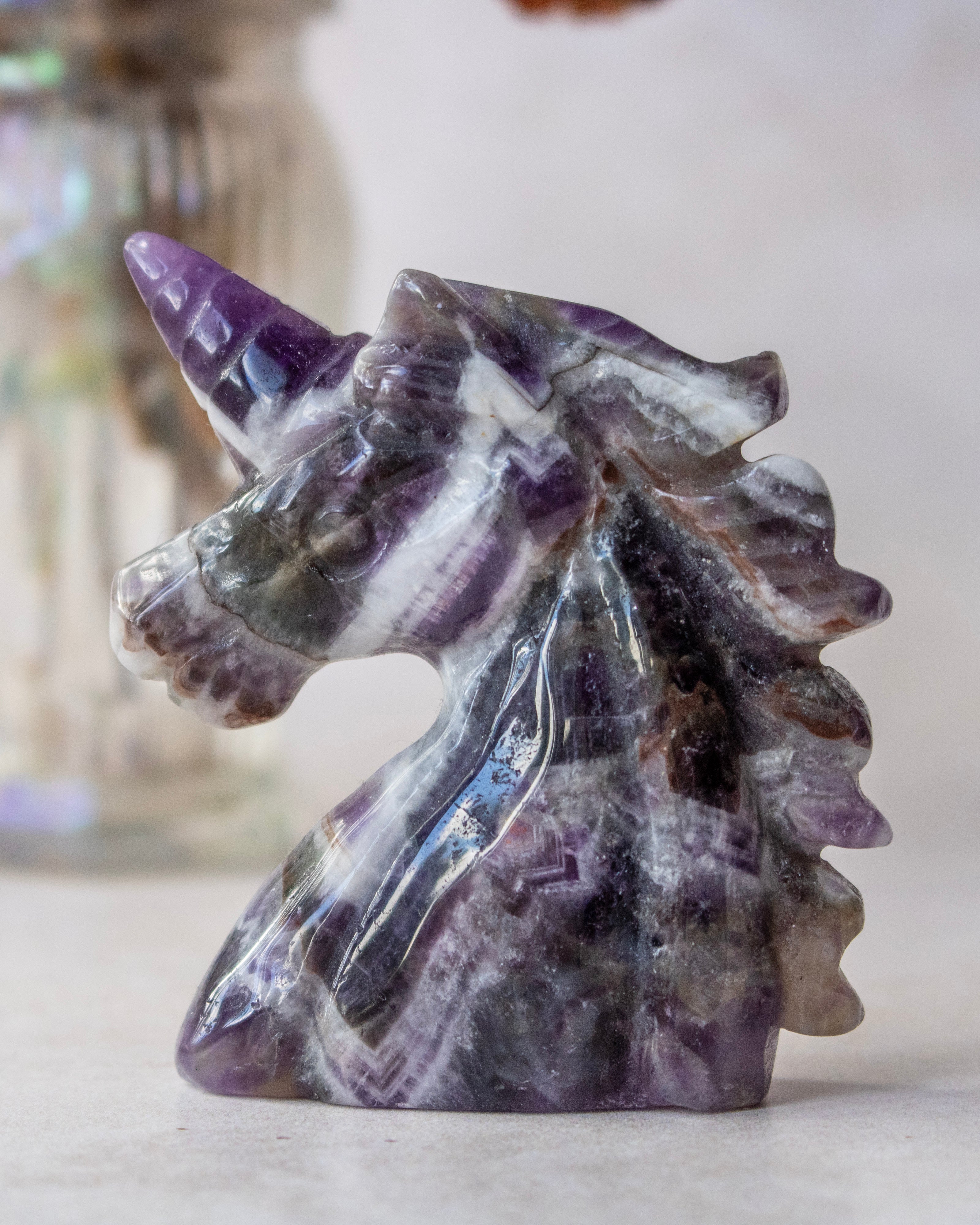 A polished purple and white striped amethyst unicorn head carving, shown in a close-up studio shot with a softly blurred background.
