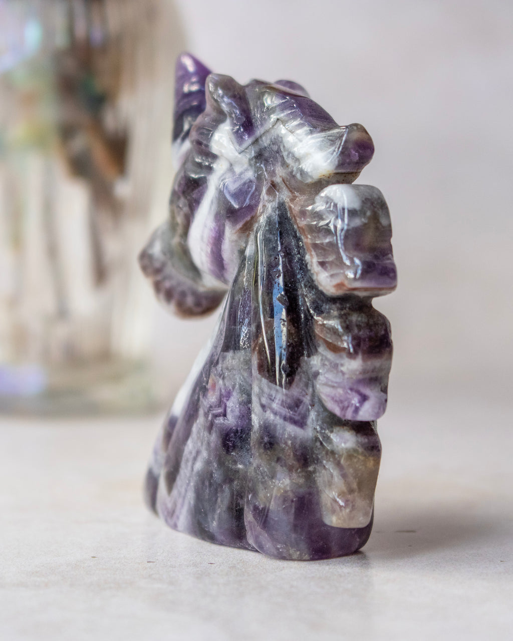 A small carved crystal figurine of a horse, made from purple and white banded fluorite, standing on a light surface with a soft-focus background.
