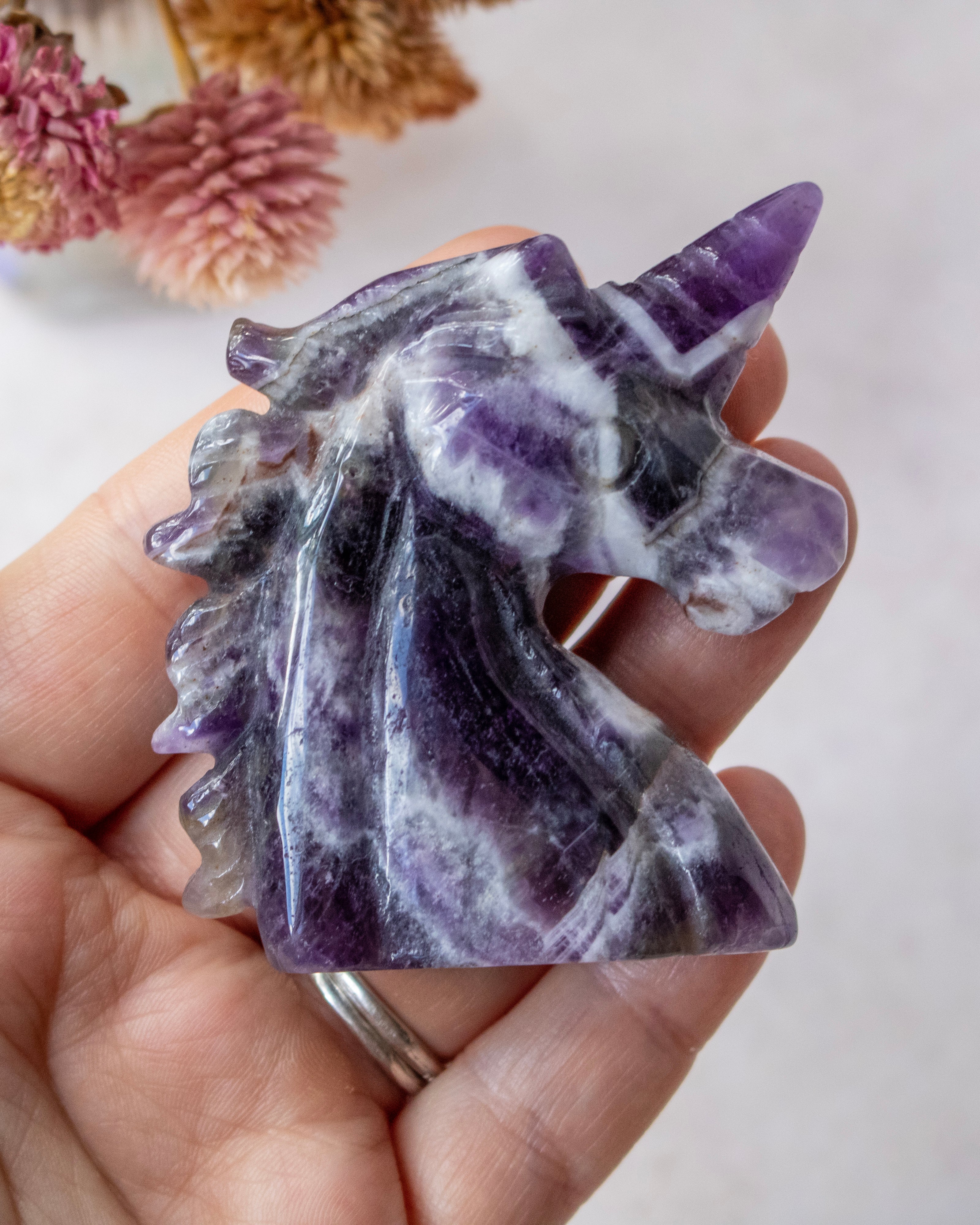 A hand holding a polished purple and white fluorite unicorn carving, with dried flowers blurred in the background.