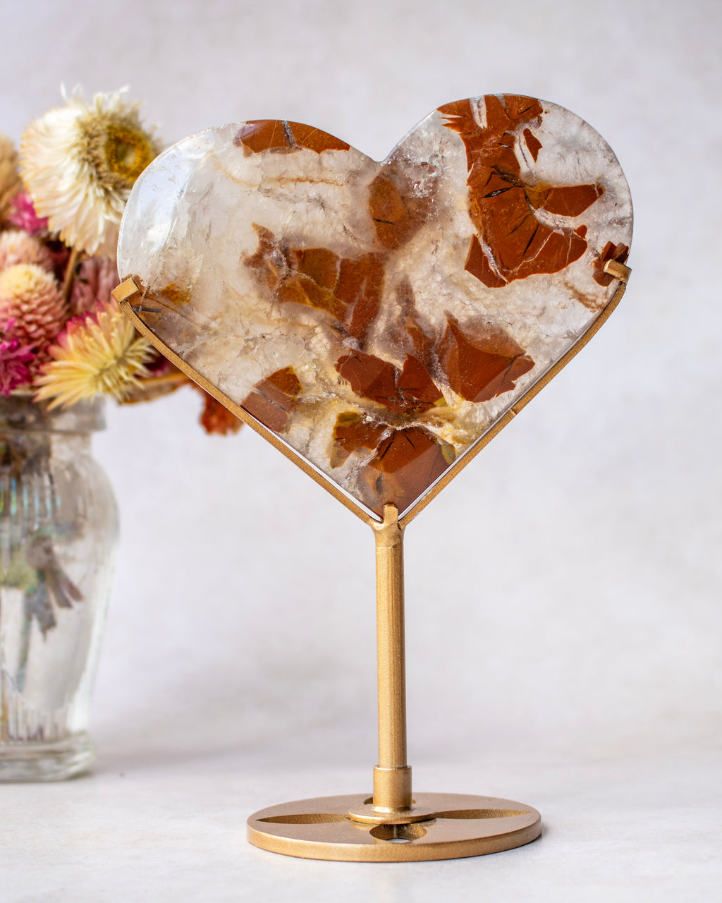 Heart-shaped crystal stone with a gold metal stand, set against a neutral light gray background with dried flowers in a glass vase on the left.