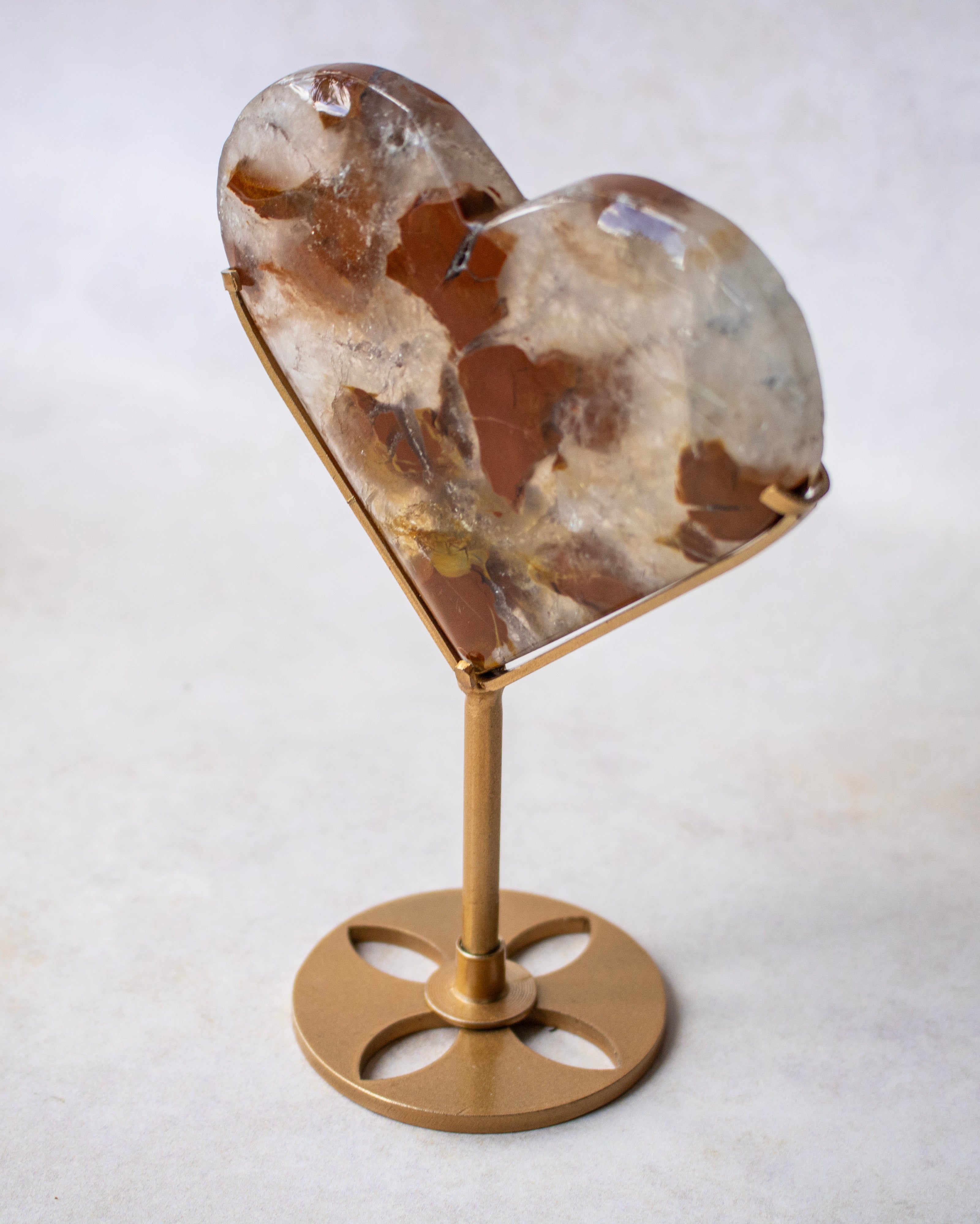 A heart-shaped crystal or stone display piece with a marbled pattern of brown, beige, and white, mounted on a gold-colored metal stand with a circular base.