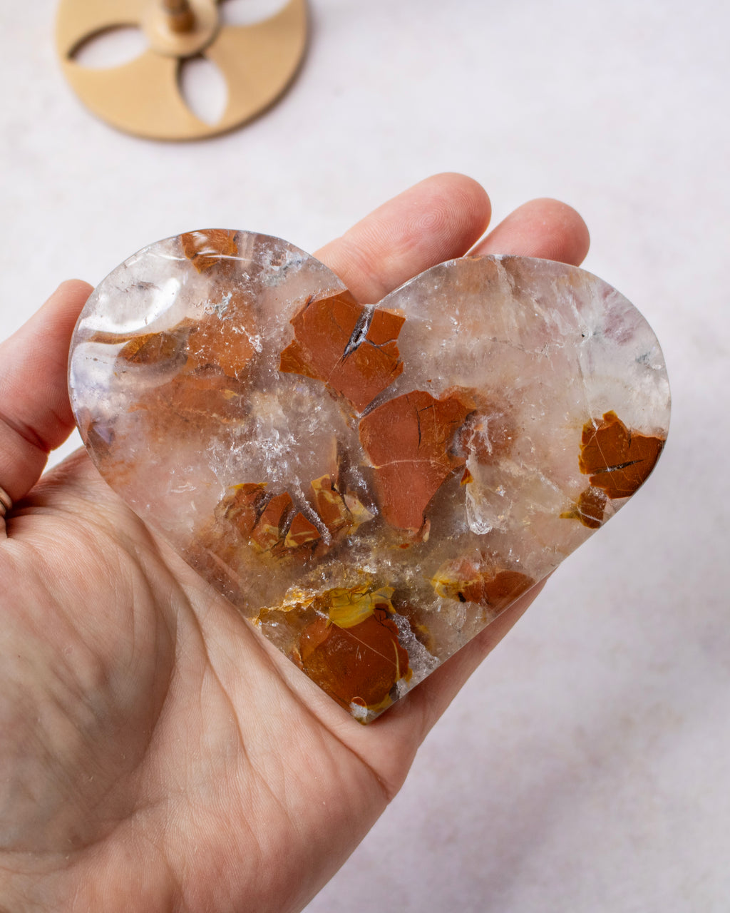 A heart-shaped polished stone or crystal held in a person's hand, showing a translucent clear body with embedded brown and golden-brown mineral inclusions. A small gold-toned circular object is partially visible in the blurred background.