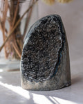 A raw crystal geode specimen with a dark gray to black crystalline interior and a silver-gray outer matrix, standing upright on a light surface with soft shadows.