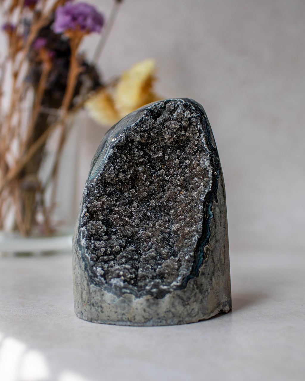 A natural black geode or crystal cluster specimen with a rough dark outer surface and a glittering black-and-gray interior, photographed on a light neutral surface with a blurred vase of dried flowers in the background.