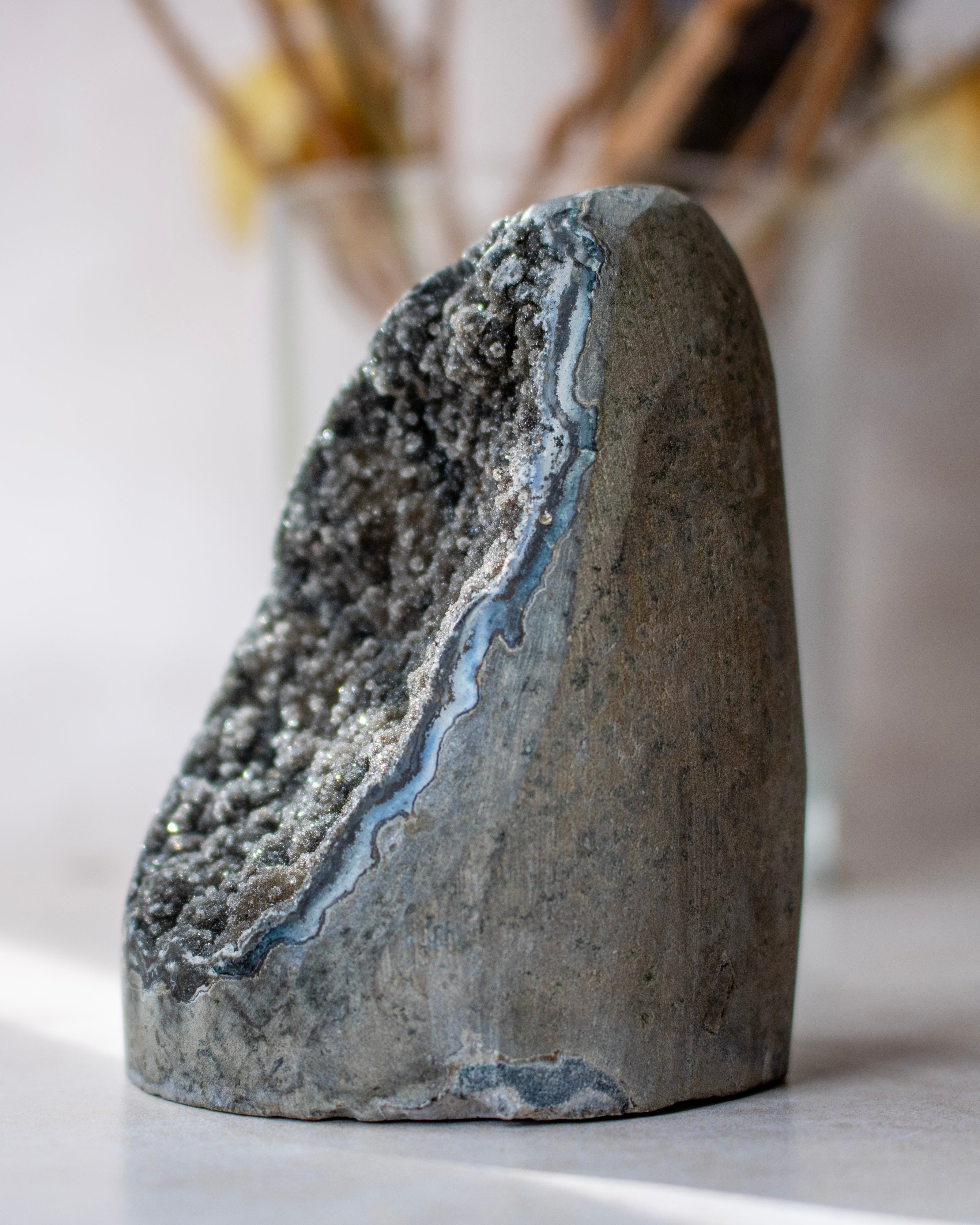 A natural geode specimen with a rough gray outer matrix and a glittering dark interior, photographed on a light surface with a softly blurred indoor background.