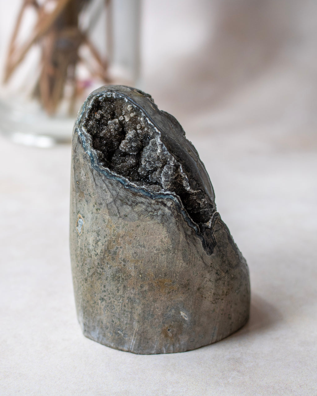 A natural gray geode specimen with a polished outer shell and a dark, crystalline interior, displayed on a neutral light beige surface.