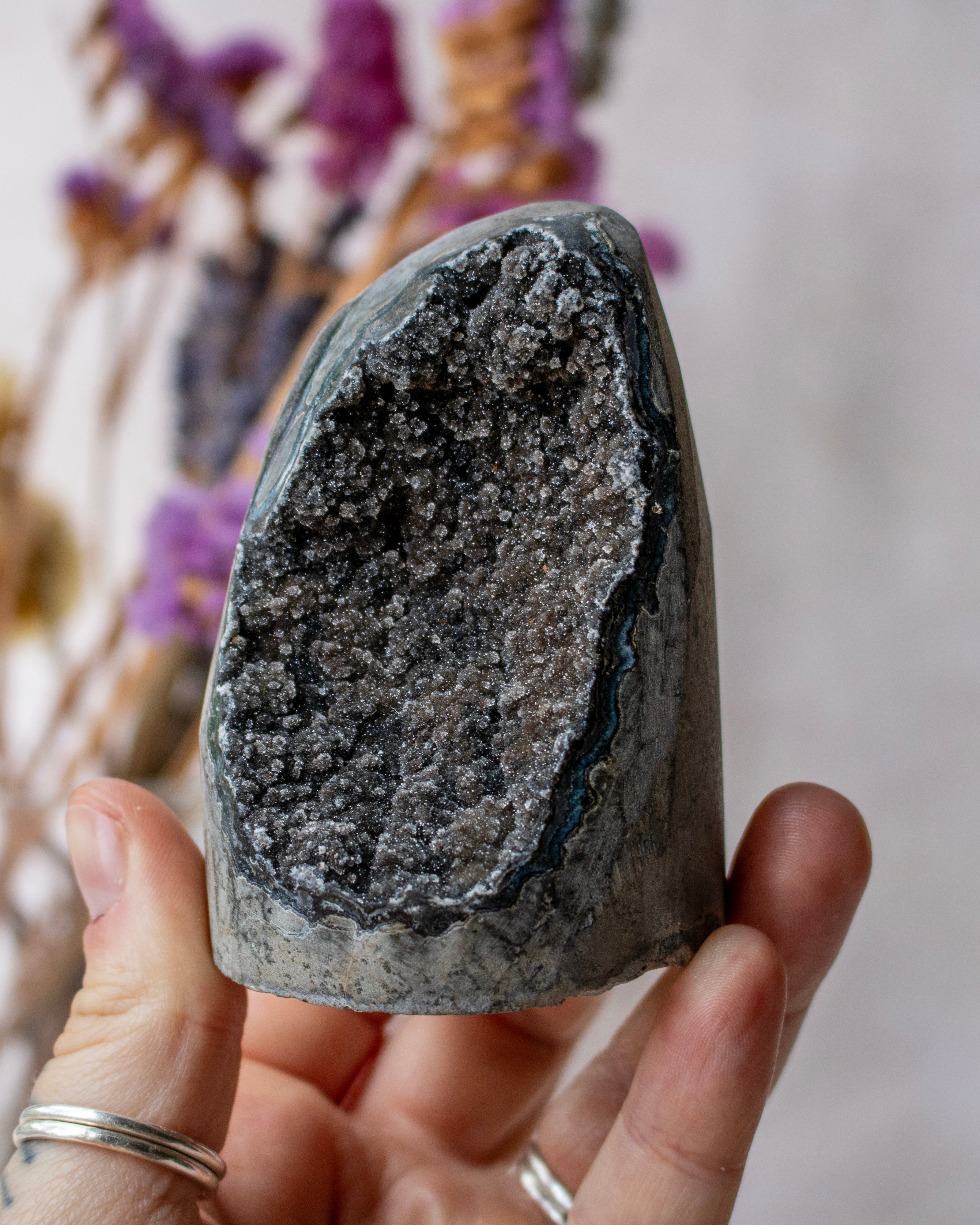 A hand holding a polished natural crystal geode with a dark gray outer matrix and a sparkling silver-gray crystalline interior, set against a soft-focus background of dried purple flowers.