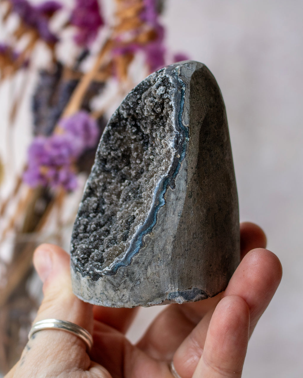 A person's hand holding a raw geode specimen with a dark gray crystalline outer layer and a bright blue inner cavity, with blurred dried purple flowers in the background.