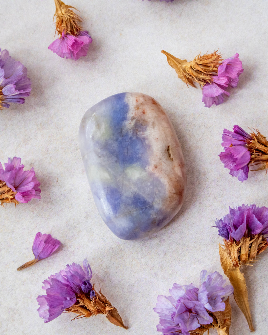 A polished tumbled stone with marbled blue, lavender, and beige patterns, styled on a textured white surface and surrounded by small dried purple flowers.