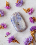 A polished lavender-blue tumbled stone crystal with natural white and gray marbling, arranged on a textured off-white surface and surrounded by dried purple flowers.