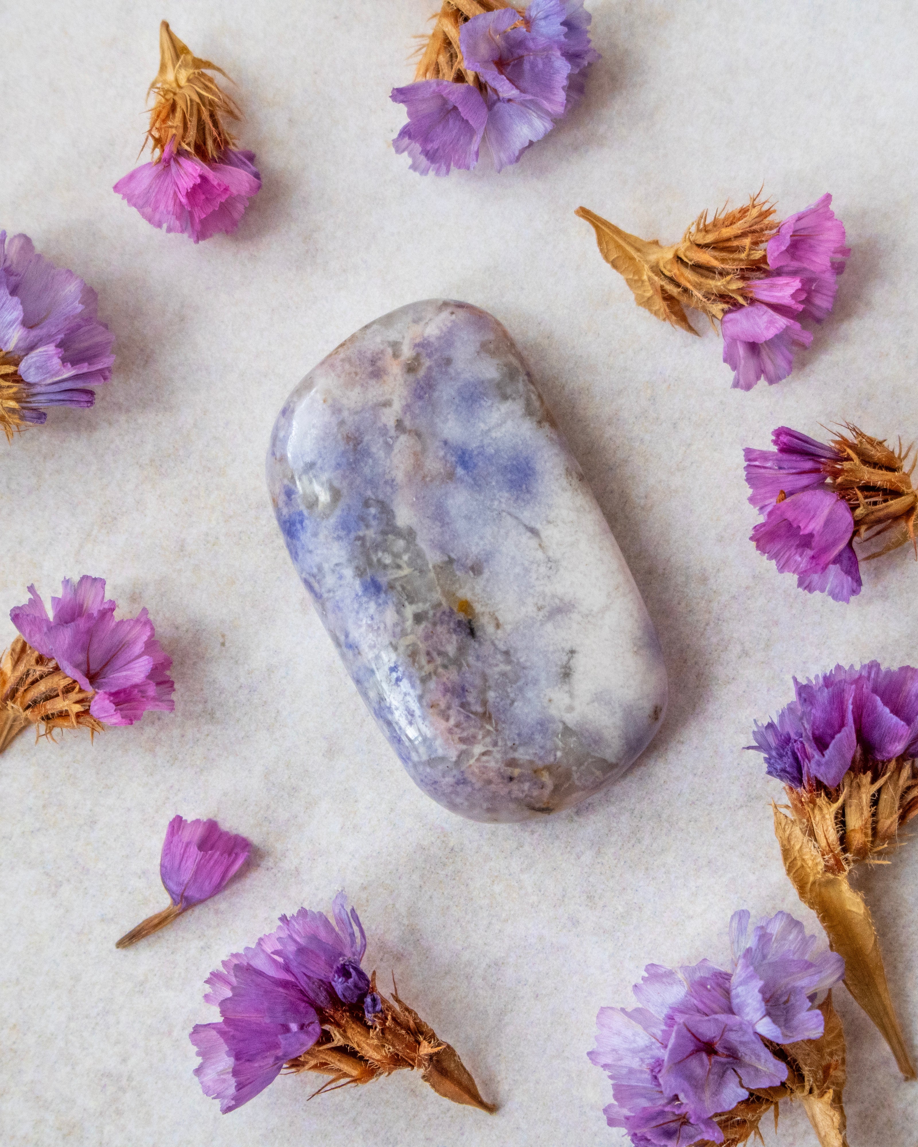 A polished lavender-blue tumbled stone crystal with natural white and gray marbling, arranged on a textured off-white surface and surrounded by dried purple flowers.