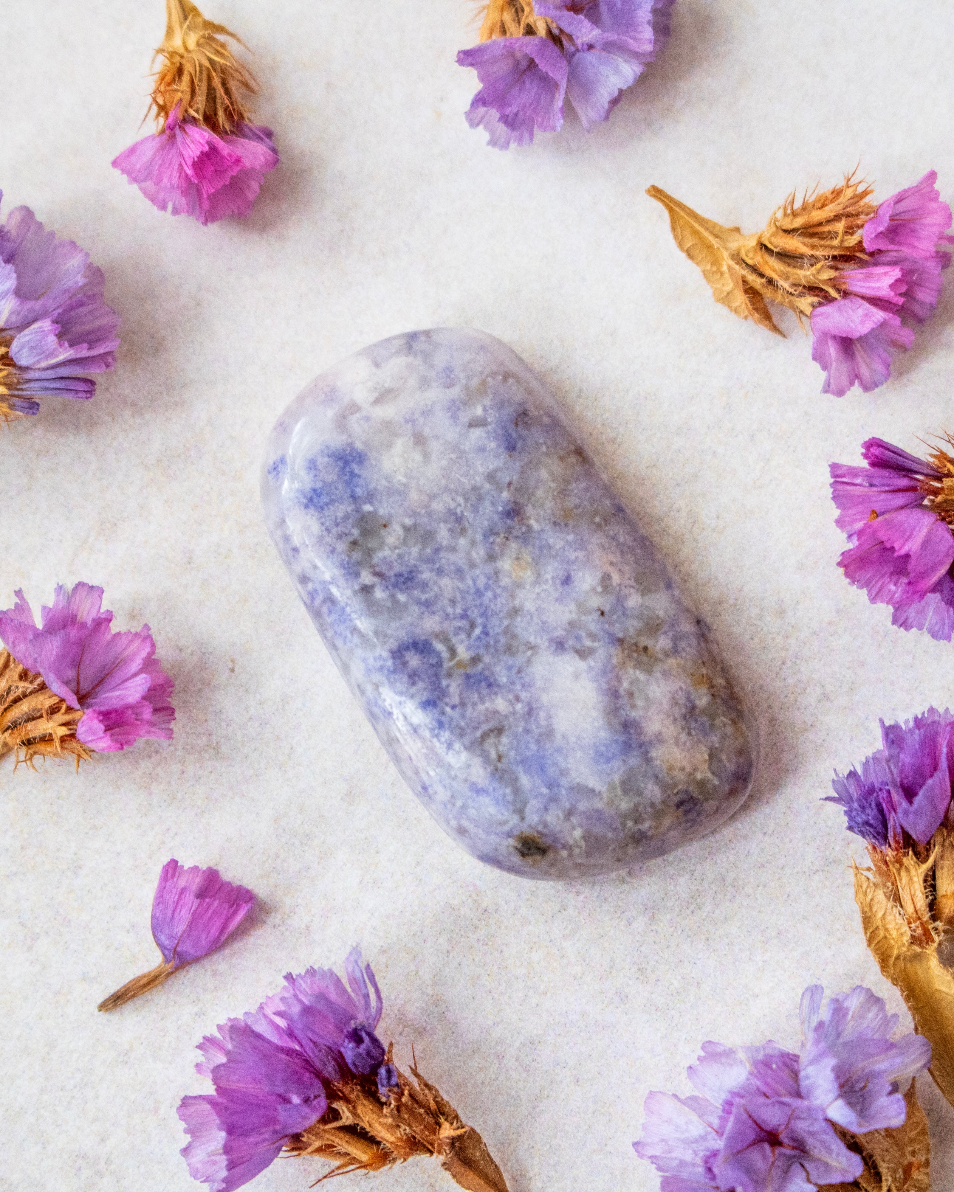 A polished lavender-blue tumbled stone crystal with natural white and gray marbling, arranged on a textured off-white surface and surrounded by dried purple flowers.