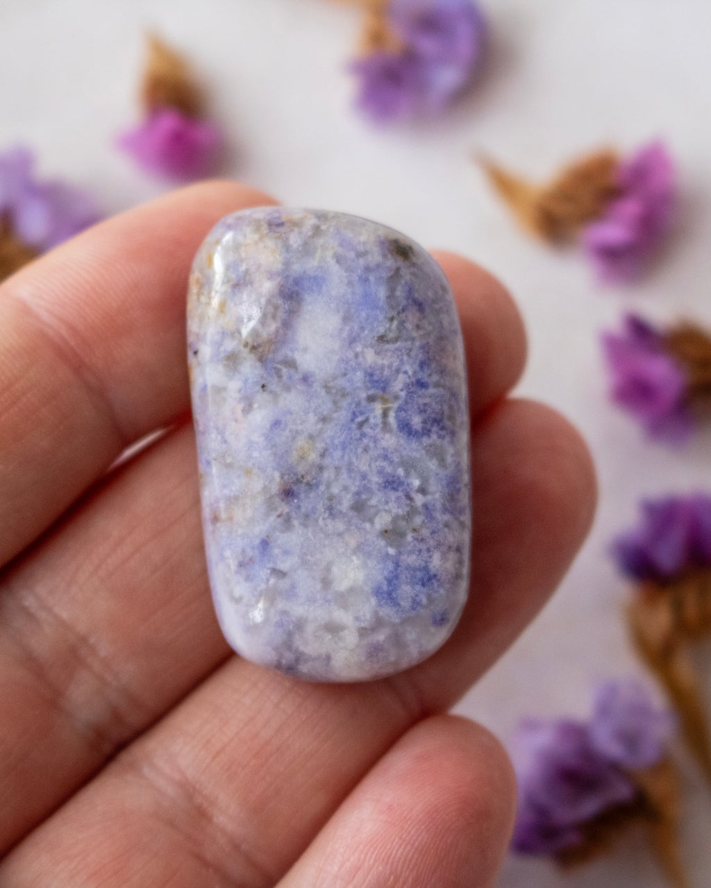 A polished lavender-blue tumbled stone crystal with natural white and gray marbling, on someone's fingers and surrounded by dried purple flowers.