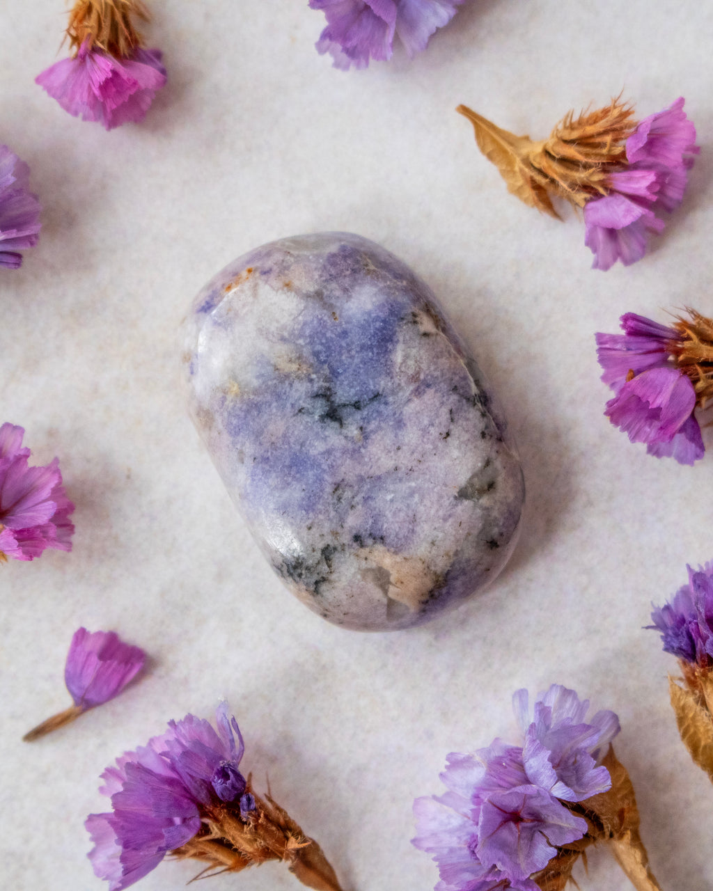 A polished lavender-blue tumbled stone crystal with natural white and gray marbling, arranged on a textured off-white surface and surrounded by dried purple flowers.